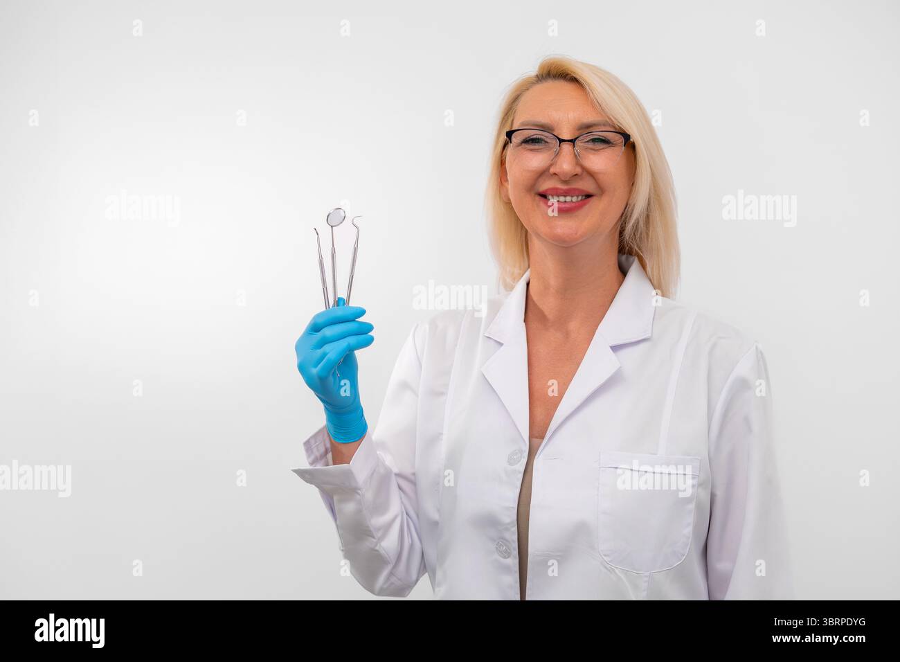 Femme dentiste blonde portant des gants et blouse de laboratoire tenant des instruments dentaires. Médecin dentaire mature en uniforme blanc avec des outils, isolé sur blanc. Concept de santé bucco-dentaire, d'hygiène et de dentisterie moderne. Banque D'Images