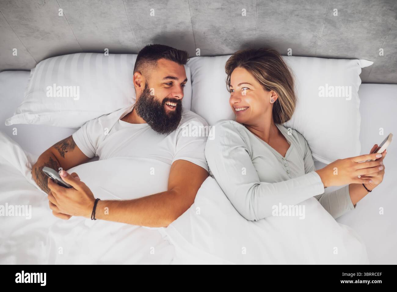 Jeune couple heureux est couché dans le lit le matin. Ils sont relaxants et utilisent des smartphones. Banque D'Images