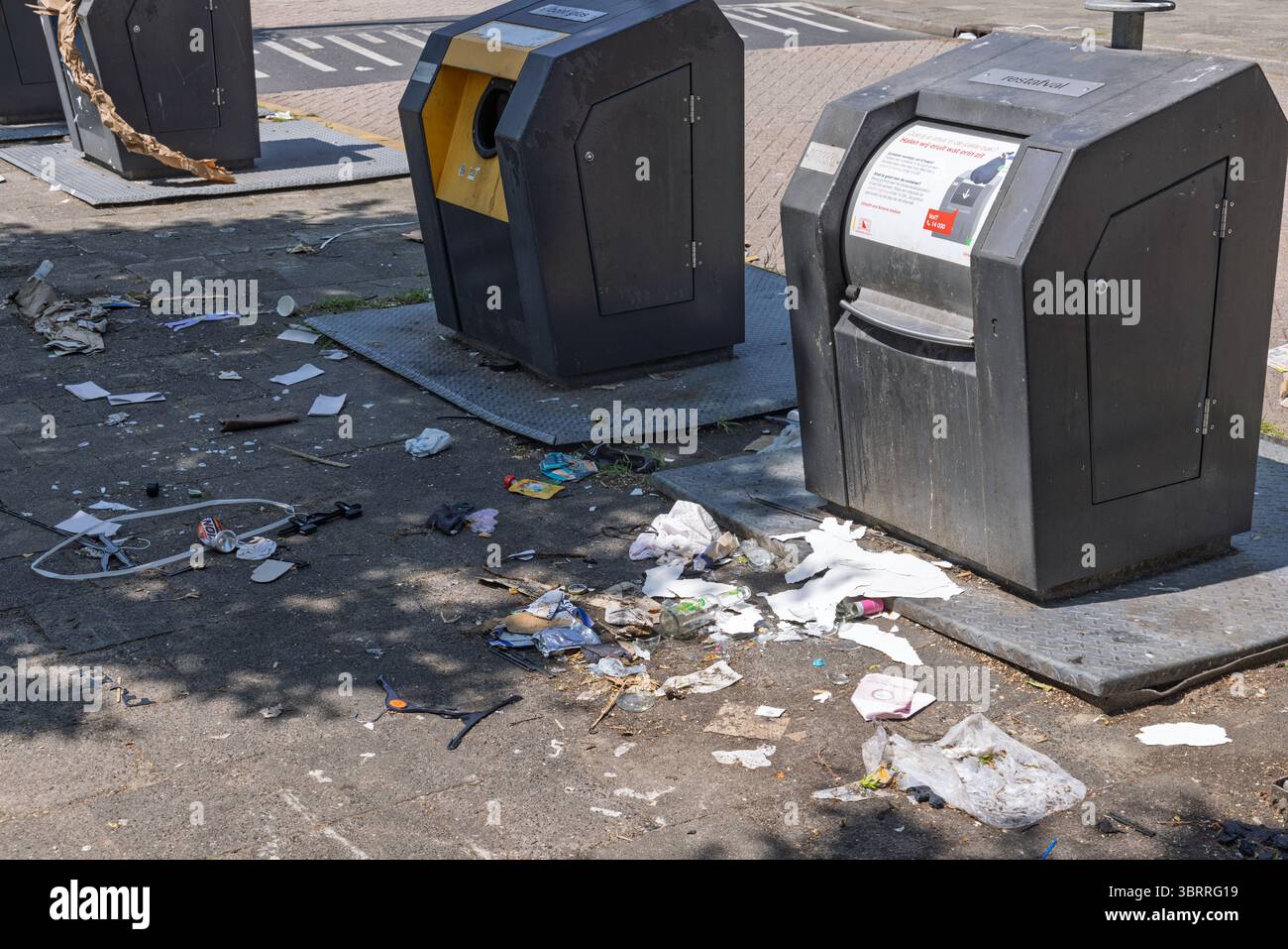 Des bacs de recyclage publics renversés entourés de déchets et de débris dispersés dans une rue ensoleillée, illustrant les défis de la gestion des déchets urbains et la pollution Banque D'Images