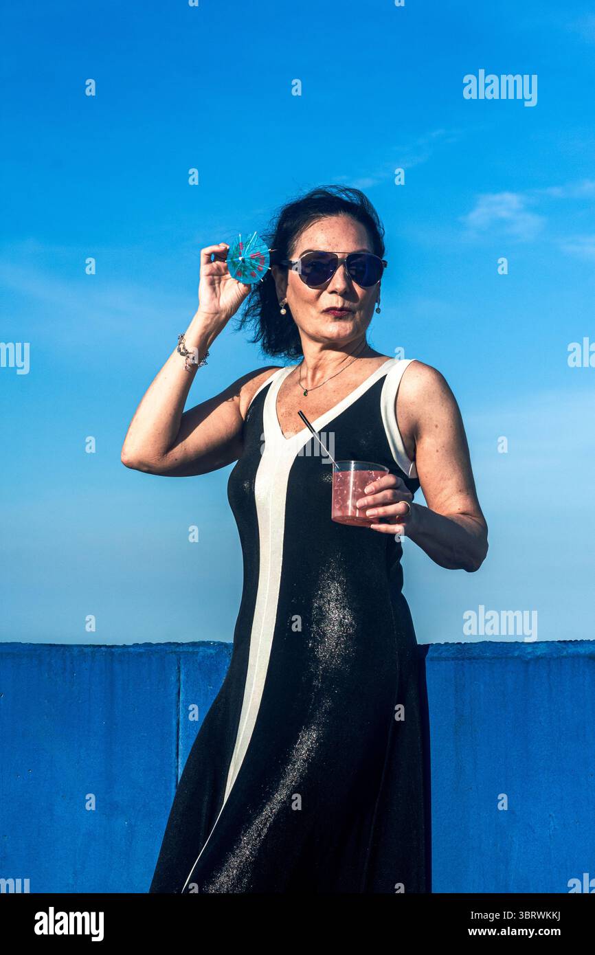 Mannequin senior élégant profitant d'un cocktail sur le toit avec un ciel bleu Banque D'Images Mannequin senior élégant profitant d'un cocktail sur le toit avec un ciel bleu Banque D'Images