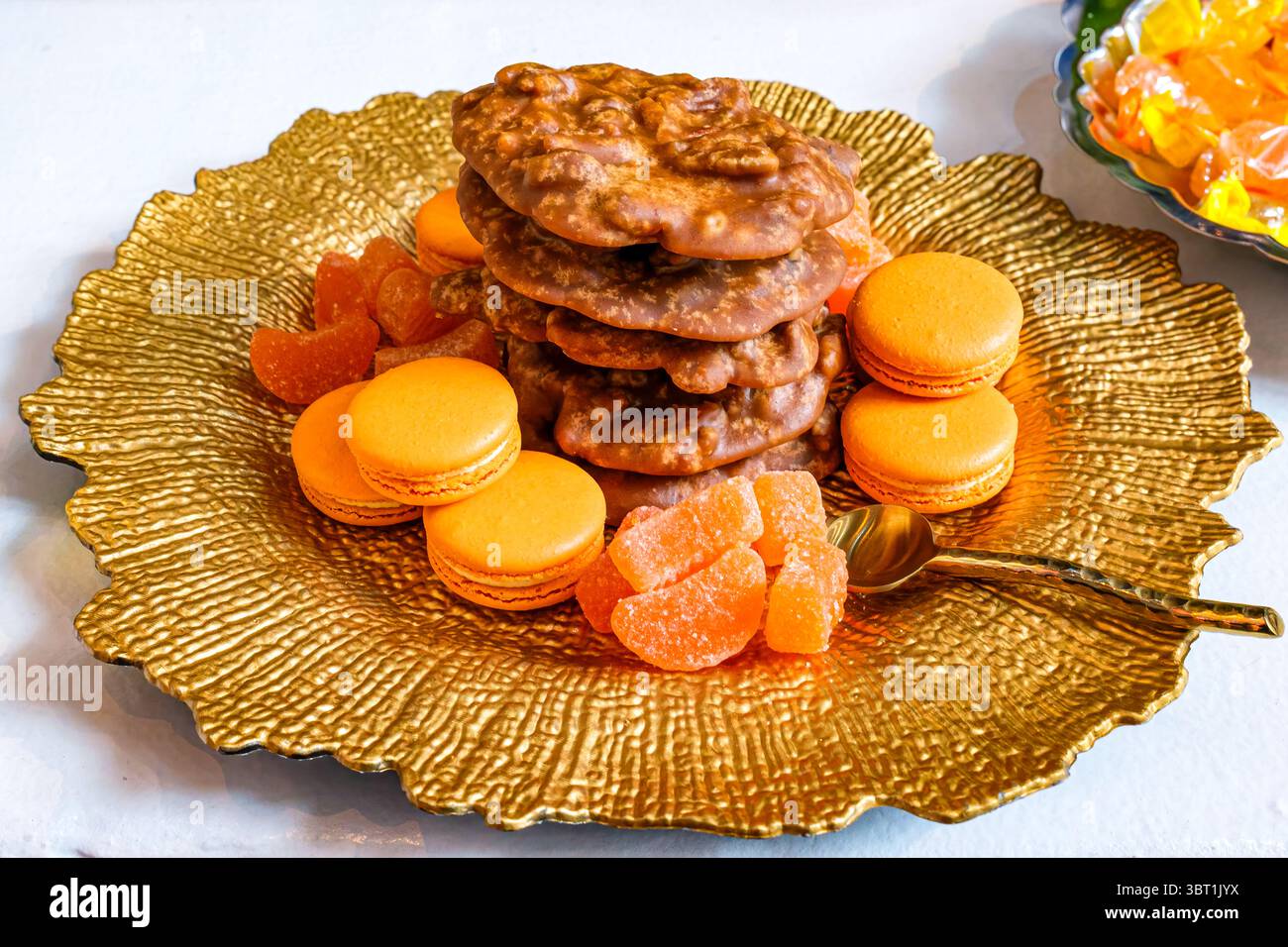 Assiette à dessert avec pile de pralines de noix de pécan, macarons et bonbons en tranches d'orange sur une plaque d'or et un fond de nappe blanche Banque D'Images