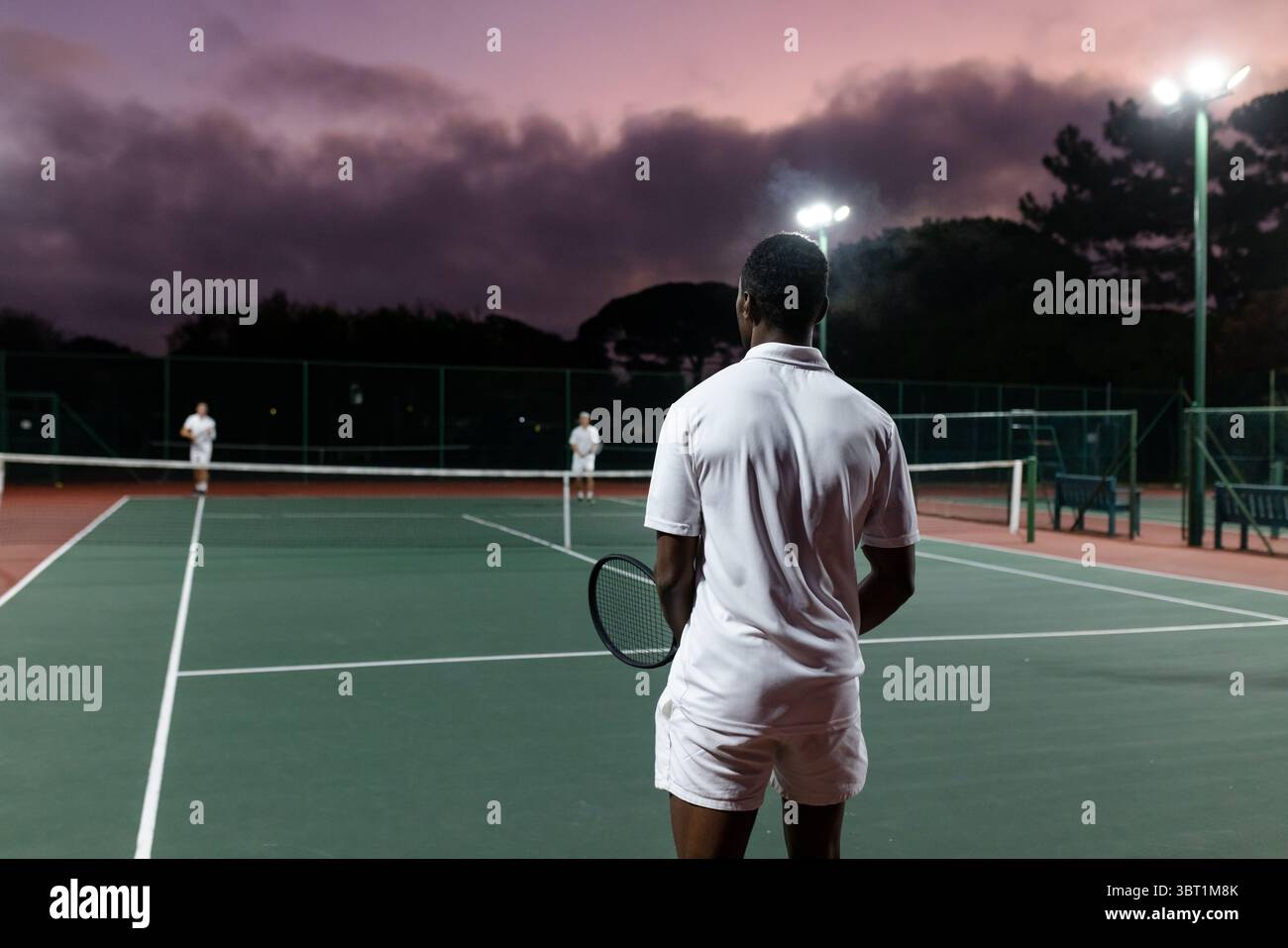 Divers joueurs de tennis masculins en compétition sur le court au crépuscule sous les projecteurs avec des raquettes, filet Banque D'Images
