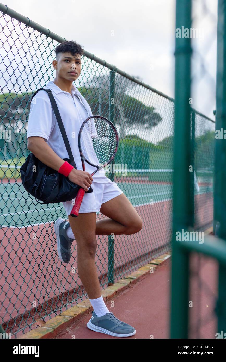 Homme afro-américain appuyé sur une clôture à maillons de chaîne sur un court de tennis tenant une raquette et un sac de sport Banque D'Images