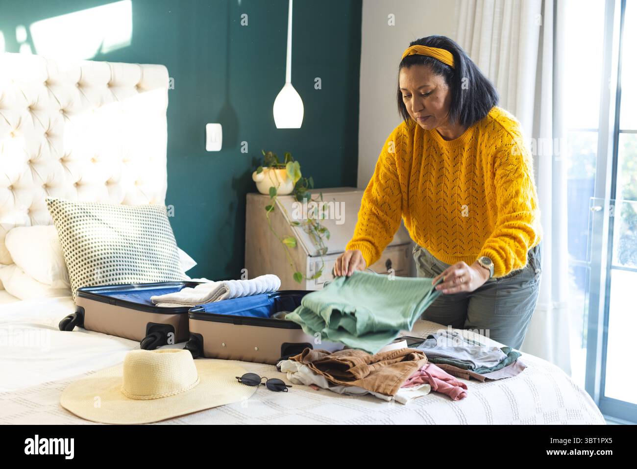 Femme pliant les vêtements de la valise ouverte à côté du lit dans la chambre avec mur d'accent vert, espace de copie Banque D'Images