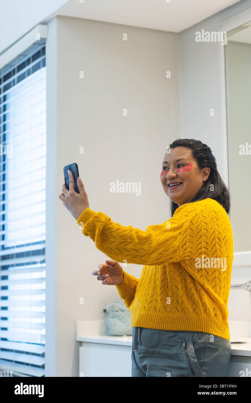 Femme adulte mature prenant selfie smartphone tout en appliquant des patchs sous les yeux dans la salle de bain Banque D'Images