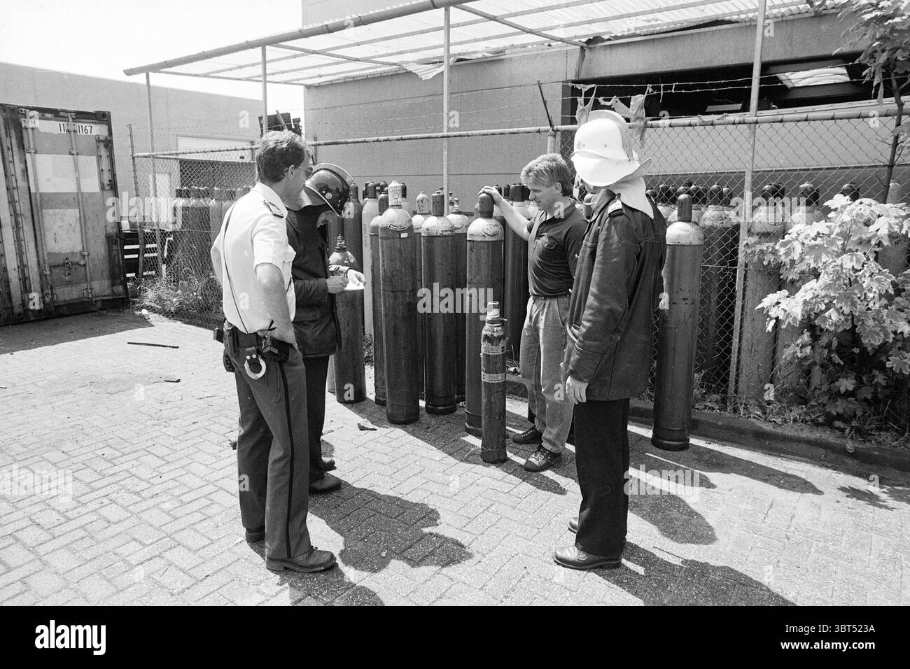 Explosion de la société de bouteilles de dioxyde de carbone R. Peereboomweg explosions Haarlem Robert Peereboomweg pays-Bas, Whizgle News, Dutch Desk, pays-Bas, 1950 - 2000 on 06-07-1987. L'image contient ces rubriques. La scène représente un groupe de quatre individus rassemblés autour d'une série de grands cylindres en métal foncé disposés en rangée. Ces bouteilles, probablement des réservoirs d'essence, dominent le premier plan, chacune montrant des signes d'usure avec des taches de peinture décolorée et des imperfections de surface. Les gens se tiennent étroitement, formant un demi-cercle autour des réservoirs, suggérant un moment de discussion ou d'inspection. Un ind Banque D'Images