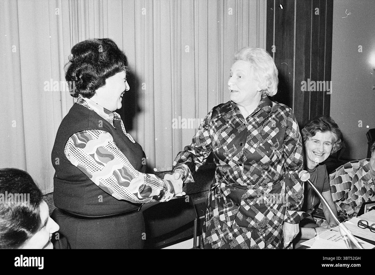 Rencontre Félicitation femme, Whizgle News, Dutch Desk, pays-Bas, 1950 - 2000 le 19-02-1974. L'image montre ces rubriques. La scène présente un rassemblement de plusieurs individus engagés dans un moment de poignée de main et d'interaction, dégageant un sentiment de camaraderie et de reconnaissance. Au premier plan, deux femmes sont au centre de l'action, échangeant une poignée de main ferme, indiquant une connexion chaleureuse. Une femme arbore une blouse à motifs caractérisée par des formes vibrantes et tourbillonnantes et des couleurs vives, suggérant les tendances de la mode de l'époque. Ses cheveux foncés encadrent son visage proprement, et son expression est o Banque D'Images