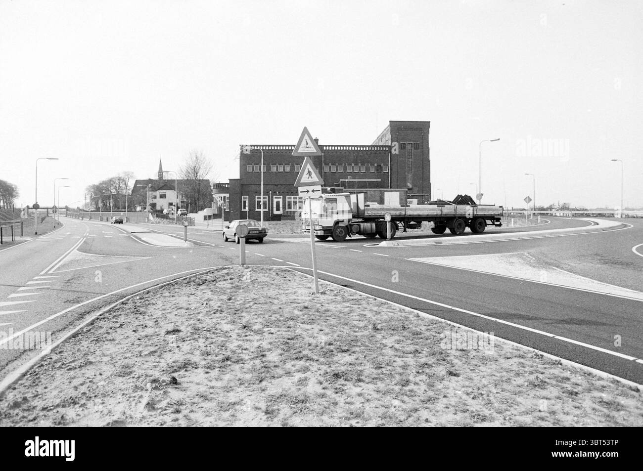 Routes Kanaaldijk (virage à droite) - de Geul IJmuiden de Geul pays-Bas, Whizgle News, Dutch Desk, pays-Bas, 1950 - 2000 on 14-03-1989. L'image montre ces rubriques. La scène capture une route circulaire avec un rond-point proéminent et large en son centre. Les zones herbeuses bordent le trottoir, suggérant un mélange d'éléments urbains et ruraux. Le rond-point comporte des panneaux de signalisation triangulaires, indiquant le flux des véhicules. Sur le côté gauche, se dresse un modeste bâtiment, présentant un style architectural robuste avec des briques sombres. Son design est quelque peu industriel, avec de grandes fenêtres et Banque D'Images