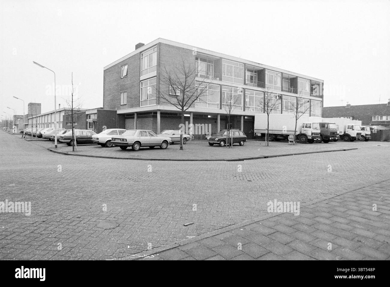 Maisons neuves Munnikenweg Beverwijk bâtiments maisons et maisons Beverwijk pays-Bas, Whizgle News, Dutch Desk, pays-Bas, 1950 - 2000 le 06-02-1981. Ce sont les éléments de l'image. La scène représente un environnement urbain sombre caractérisé par une palette principalement grise, suggérant une journée silencieuse, peut-être nuageuse. Au premier plan, une large rue pavée courbe doucement, menant le regard du spectateur vers une collection de véhicules stationnés qui bordent les deux côtés. À droite, plusieurs voitures, pour la plupart des berlines, reposent calmement le long du trottoir. Ils varient en couleur, avec certains étant des nuances plus foncées o Banque D'Images