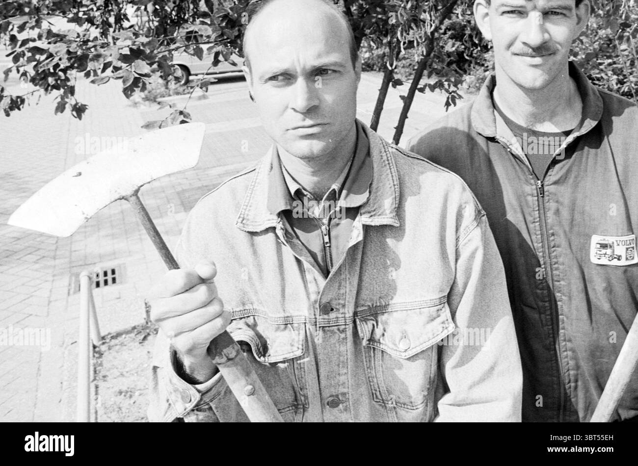 Public Gardens Service & The tunnel, Whizgle News, Dutch Desk, pays-Bas, 1950 - 2000 on 05-08-1993. L'image inclut ces rubriques. La scène montre deux hommes dans un moment franc, debout près l'un de l'autre sur fond de verdure et d'architecture urbaine. Le premier homme, bien en vue au premier plan, tient fermement dans sa main droite un outil de jardinage, un outil courbe rappelant une faucille. Il a une expression sérieuse, suggérant détermination ou concentration. Vêtu d'une veste en denim clair qui contraste avec les tons foncés de son environnement, sa tête rasée de près reflète un Banque D'Images