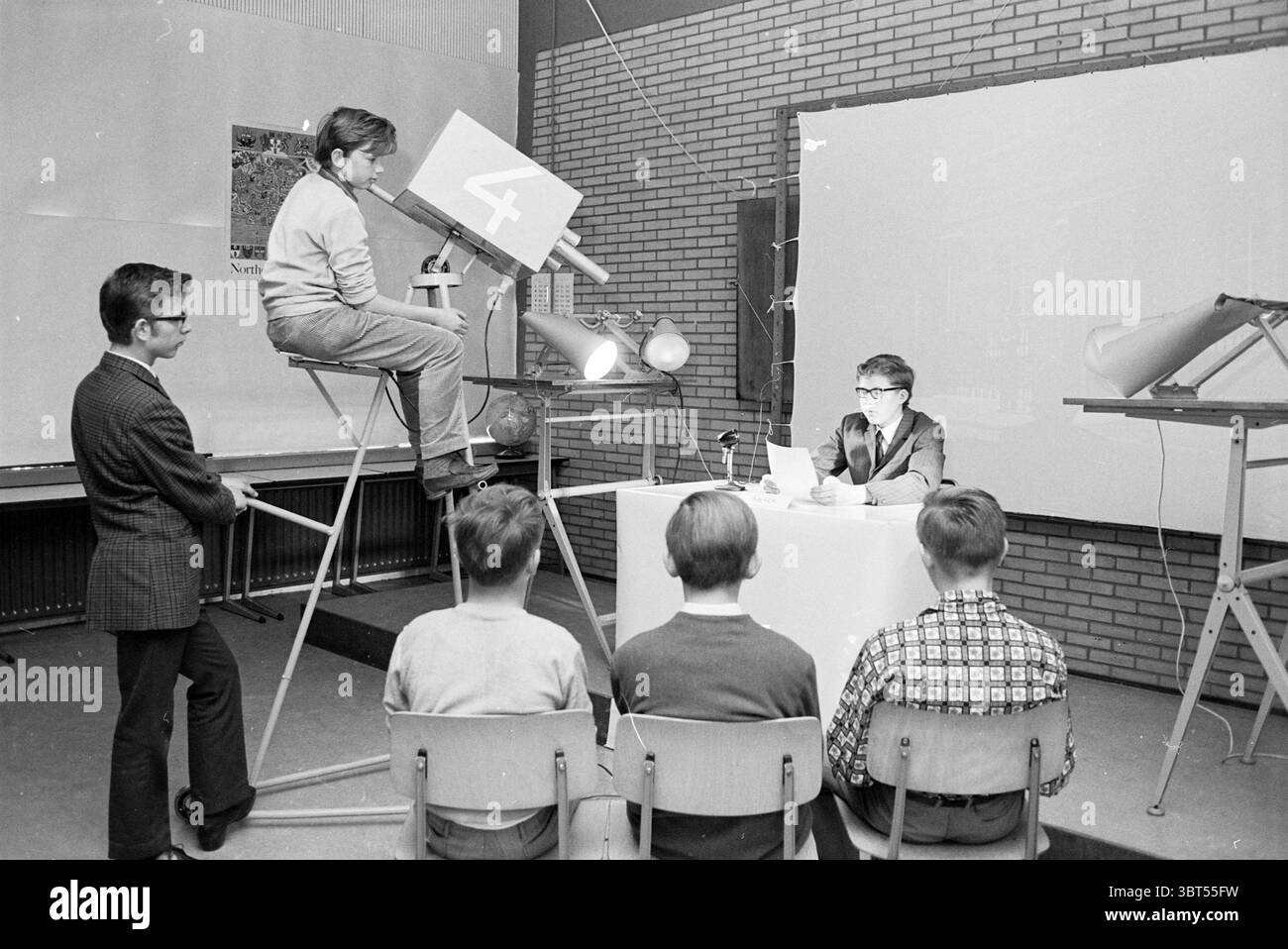 Cours d'anglais papier école de commerce écoles, Whizgle News, Dutch Desk, pays-Bas, 1950 - 2000 le 26-04-1968. L'image contient ces rubriques. Dans une salle de classe ou un studio, un groupe d'enfants est engagé dans une activité éducative ludique ressemblant à une émission de nouvelles. La chambre dispose d'une toile de fond de murs de briques texturés, ajoutant une atmosphère chaleureuse et nostalgique à l'environnement. Au premier plan, un garçon est assis derrière une petite table, lisant avec confiance dans une pile de papiers, se considérant peut-être comme un présentateur de nouvelles. Il porte des lunettes et est habillé dans une tenue élégante, exsudant un sentiment de seriousne Banque D'Images