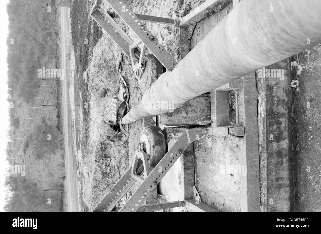Pipeline construction Hoogovens North West Hoogovens, Whizgle News, Dutch Desk, pays-Bas, 1950 - 2000 on 27-04-1986. L'image inclut ces rubriques. La scène présente un paysage industriel marqué rendu dans des tons monochromes, mettant l'accent sur les textures et les formes plutôt que sur la couleur. Au premier plan, un grand tuyau cylindrique domine la composition, sa surface montrant des signes d'usure avec une texture rugueuse qui capte la lumière, créant des contrastes et des ombres subtils. Entourant le tuyau, un cadre de supports métalliques angulaires ajoute une sensation de structure et de rigidité. Le cadre est marqué par boulon Banque D'Images