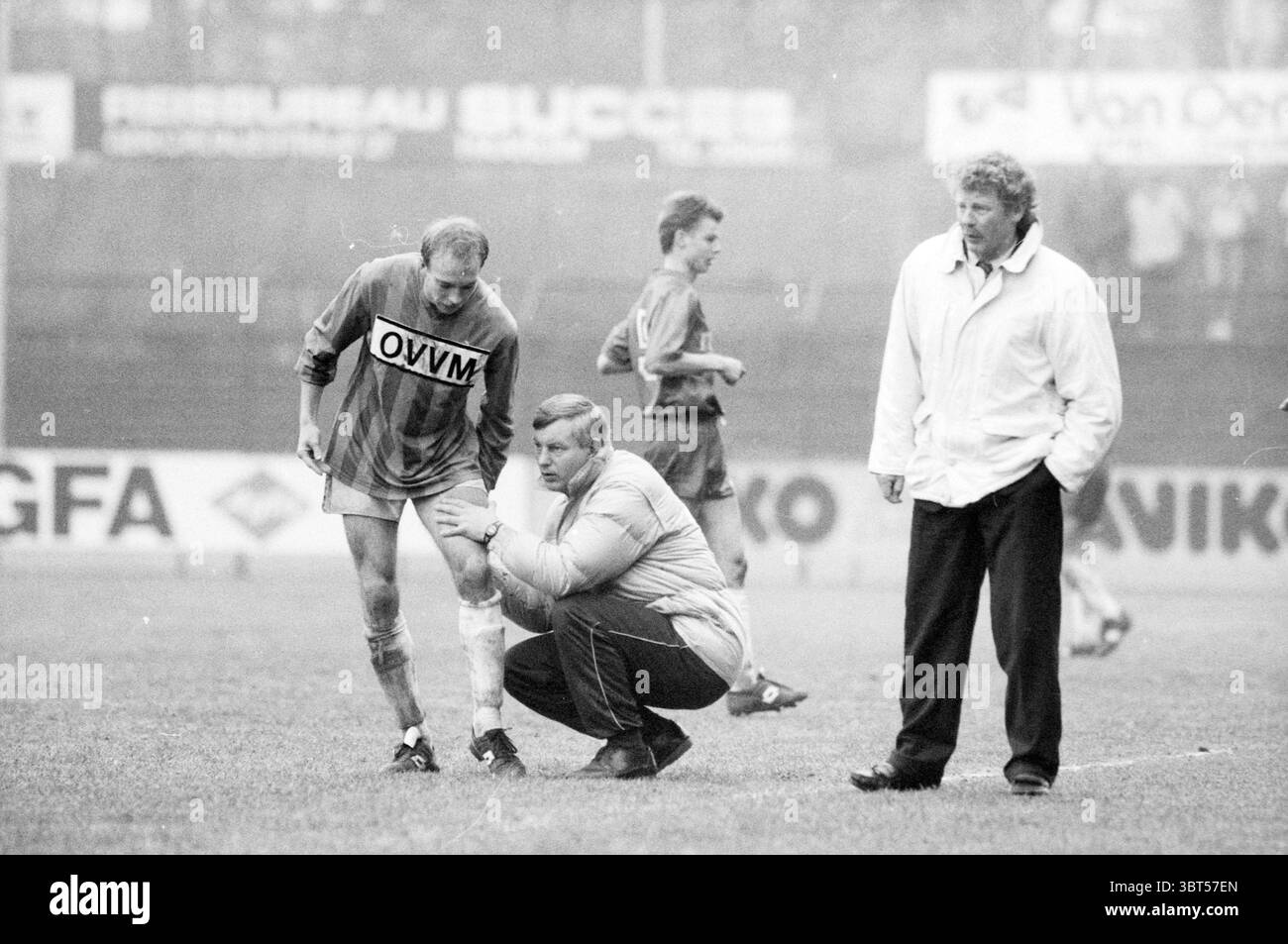 HFC Haarlem - Willem II, Whizgle News, Dutch Desk, pays-Bas, 1950 - 2000 on 03-1989. Voici les rubriques de l'image. La scène capture un moment intense lors d'un événement sportif, probablement un match de football. Le cadre est un stade en plein air, avec un terrain herbeux qui semble légèrement humide, suggérant un temps couvert ou une pluie récente. Au premier plan, deux individus sont au centre de l'attention. Un joueur, vêtu d'un maillot orange avec un logo visible, regarde son genou tandis qu'un entraîneur, agenouillé à côté de lui, examine l'état du joueur avec une expression préoccupante. L'entraîneur est vêtu de moi Banque D'Images