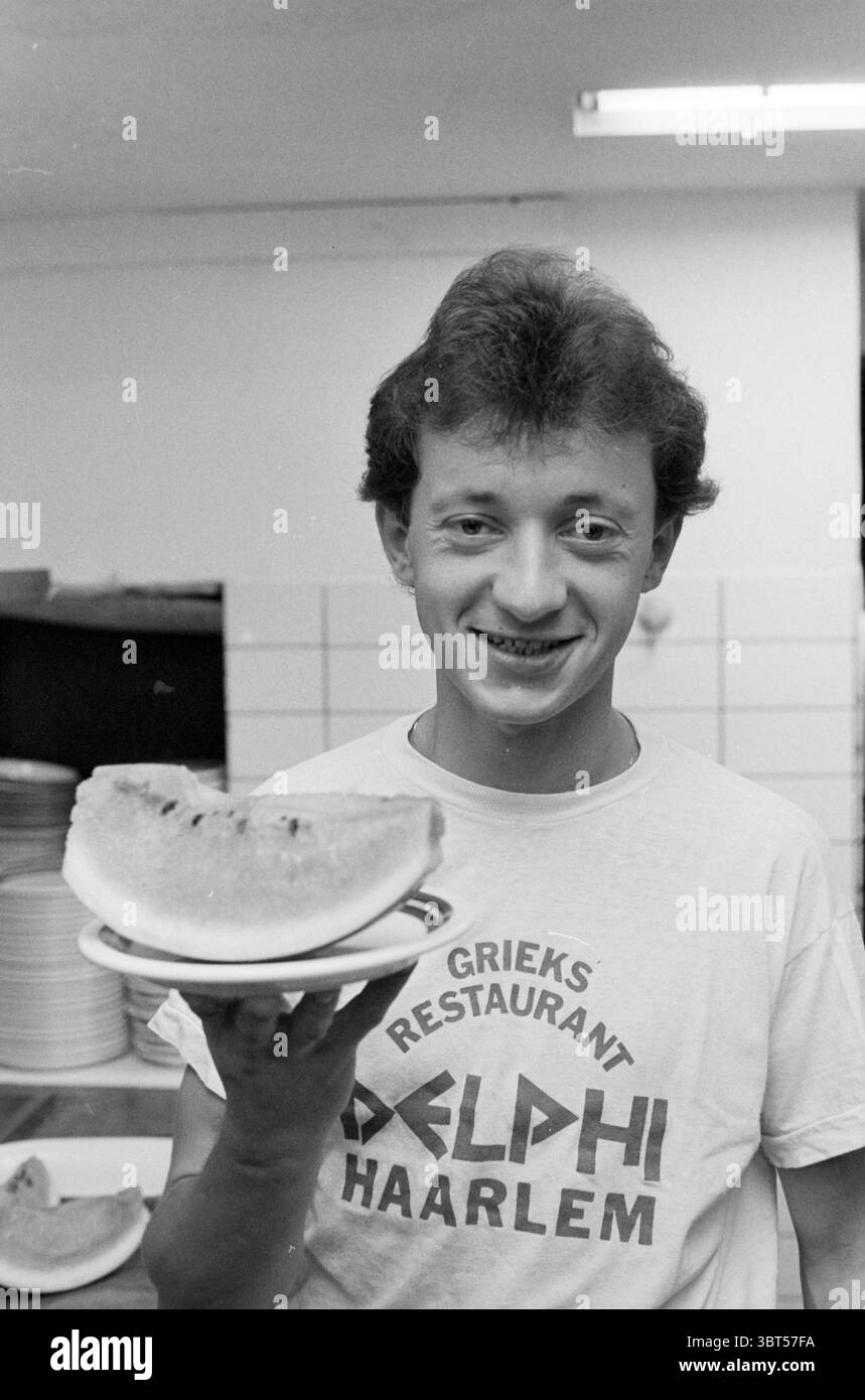 Cuisinez les Lolas REST. Delphi Gravinnesteeg Cooking Restaurant Haarlem Gravinnesteeg pays-Bas, Whizgle News, Dutch Desk, pays-Bas, 1950 - 2000 le 21-07-1987. L'image contient ces rubriques. Dans un cadre intérieur bien éclairé, un jeune se tient debout avec confiance, tenant une tranche de pastèque sur une petite assiette. L'atmosphère est décontractée et conviviale, véhiculée par le large sourire du sujet et sa posture détendue. L'individu a les cheveux courts et légèrement bouclés et arbore un T-shirt simple de couleur claire avec un nom de restaurant bien en vue sur le devant. La tranche de pastèque est vibrante, showc Banque D'Images
