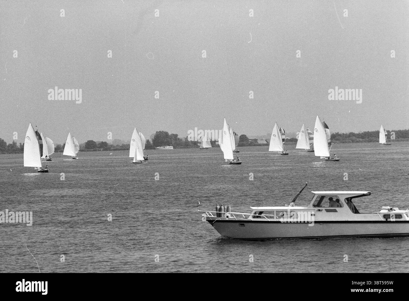 Voile : rainbow class Braasemermeer voile, Whizgle News, Dutch Desk, pays-Bas, 1950 - 2000 le 05-07-1979. Voici les rubriques de l'image. La scène capture un cadre serein en bord de mer, où une multitude de voiliers glissent gracieusement sur un plan d'eau calme. Chaque voilier a sa voile blanche déployée, attrapant la brise, tandis que leurs coques flottent doucement sur la surface, créant des ondulations subtiles. Les voiles varient légèrement en taille et en forme, contribuant à un modèle rythmé à travers l'eau. Au premier plan, un plus grand bateau à moteur se déplace régulièrement, son corps blanc et élégant contrastant avec le th Banque D'Images