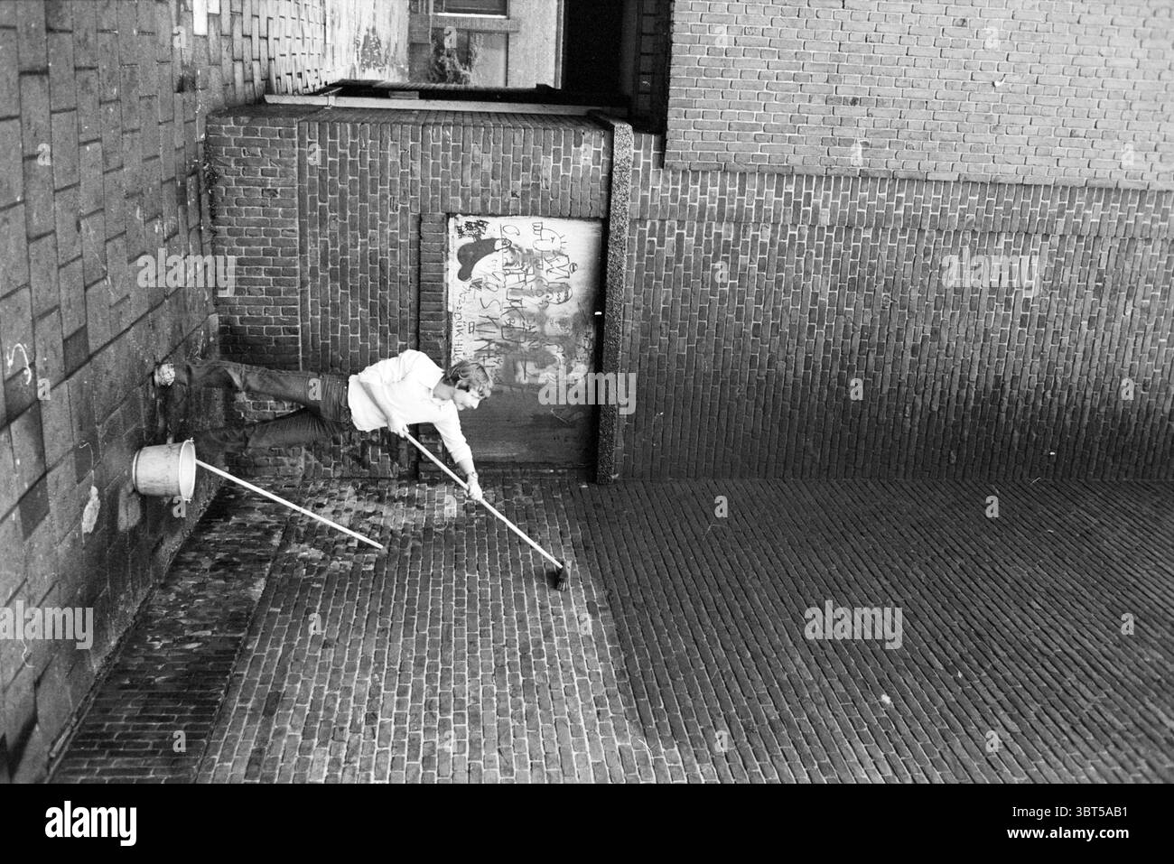 Nettoyeur scolaire Karolingenstraat Ecoles Haarlem Karolingenstraat pays-Bas, Whizgle News, Dutch Desk, pays-Bas, 1950 - 2000 on 17-11-1981. L'image montre ces rubriques. Dans une ruelle urbaine étroite, le sol est pavé de briques altérées, créant une texture rustique. Les murs environnants, construits à partir de briques rouge foncé et brun, portent des signes d'âge avec des taches de peinture écaillée et des graffitis pâles, donnant une impression de grain à la scène. Au premier plan, une figure en tenue décontractée se penche en avant, en utilisant un balai pour balayer le sol. L'individu est concentré sur sa tâche et son postur Banque D'Images