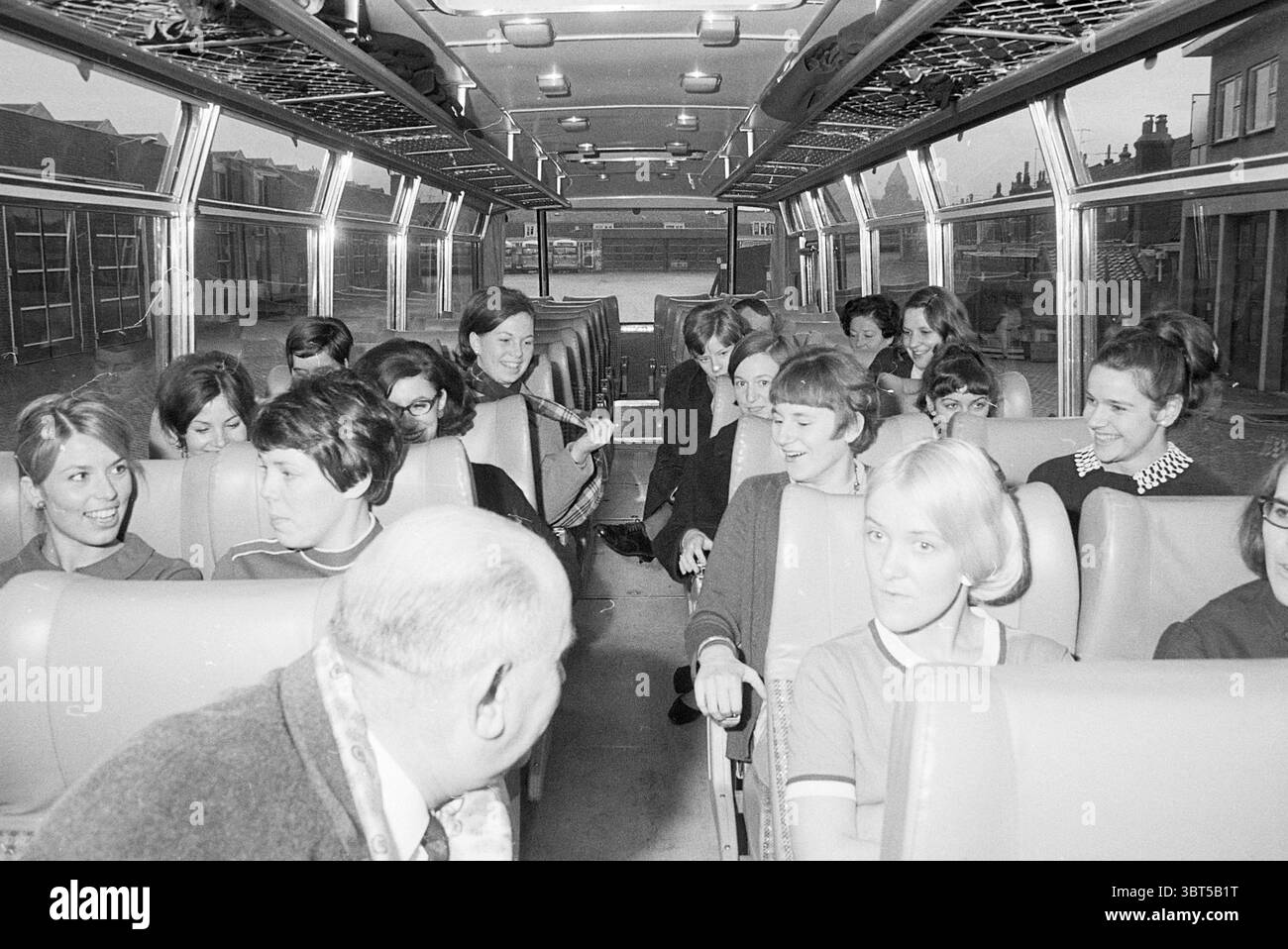Visitez NS informateurs à NZH Noord Zuid Hollandse Vervoersmaatschappij N.V. NZ, Whizgle News, Dutch Desk, pays-Bas, 1950 - 2000 le 16-02-1968. L'image montre ces rubriques. La scène représente l'intérieur d'un bus rempli d'un groupe de jeunes femmes et de quelques individus plus âgés. Le bus a un design spacieux et moderne pour son époque, avec de longues rangées de sièges de couleur claire avec une texture douce et rembourrée. La disposition des sièges est quelque peu informelle, créant une atmosphère chaleureuse parmi les passagers. Les jeunes femmes sont habillées dans une variété de tenues typiques des années 1960, caractérisées par bo Banque D'Images