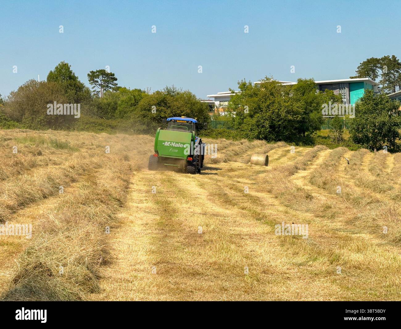 Pontypridd, pays de Galles, Royaume-Uni - 12 juillet 2025 : tracteur remorquant une machine de pressage pour fabriquer de grandes balles rondes à partir d'herbe séchée dans un champ agricole. - Image de stock capturée avec un smartphone