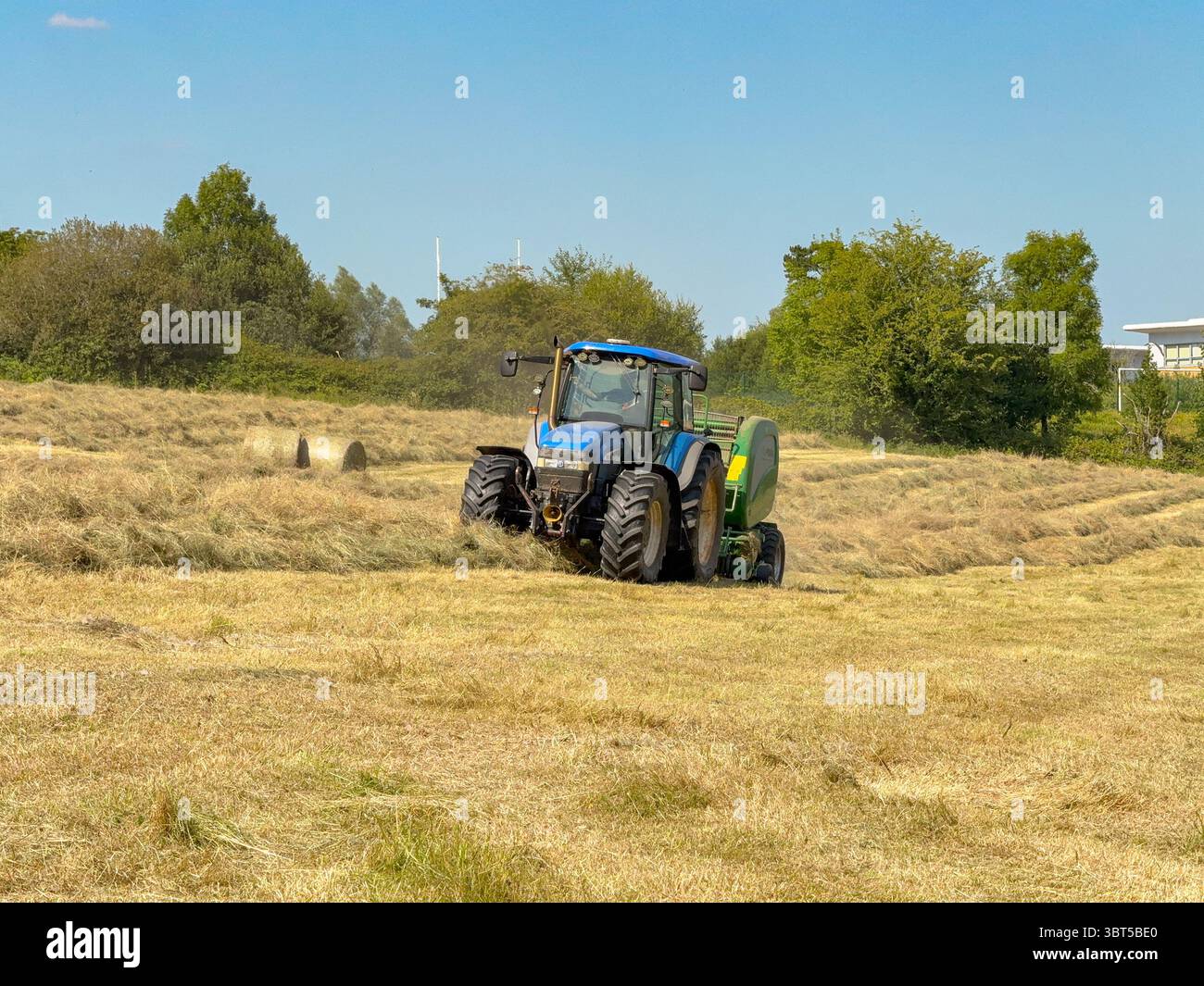 Pontypridd, pays de Galles, Royaume-Uni - 12 juillet 2025 : tracteur remorquant une machine de pressage pour fabriquer de grandes balles rondes à partir d'herbe séchée dans un champ agricole. - Image de stock capturée avec un smartphone