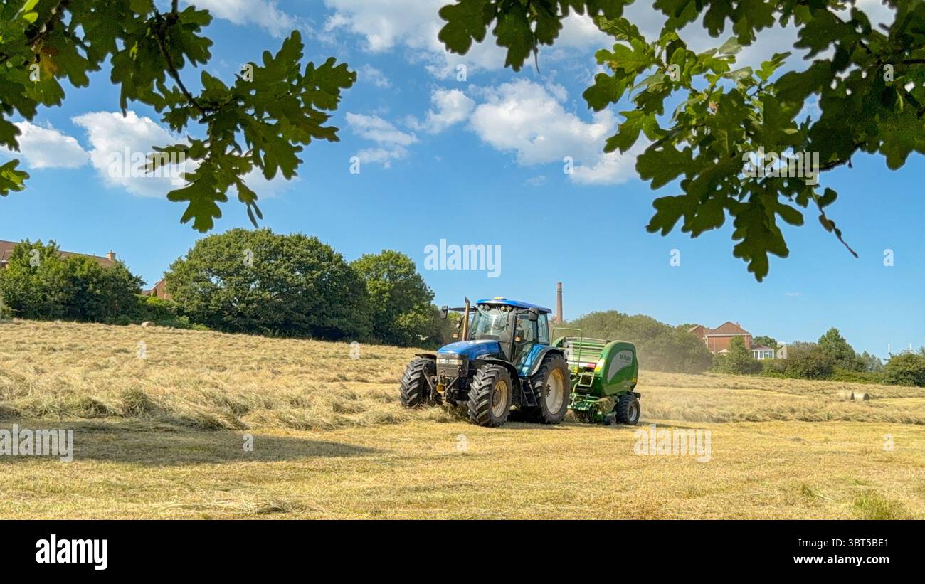 Pontypridd, pays de Galles, Royaume-Uni - 12 juillet 2025 : tracteur remorquant une machine de pressage pour fabriquer de grandes balles rondes à partir d'herbe séchée dans un champ agricole. - Image de stock capturée avec un smartphone