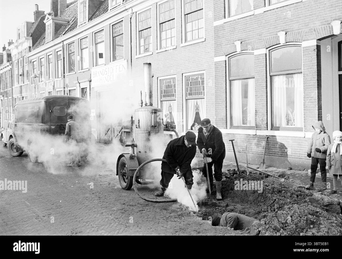 Réparation de conduites d'eau travaux de conduites d'eau, Whizgle News, Dutch Desk, pays-Bas, 1950 - 2000 on 20-01-1963. L'image montre ces rubriques. La scène représente une rue animée dans une ville caractérisée par des bâtiments en briques avec de grandes fenêtres et des détails architecturaux classiques. Les bâtiments sont disposés de manière linéaire, présentant une gamme de couleurs sourdes telles que les bruns doux, les blancs et les tons pastel, faisant allusion à un contexte historique. Au premier plan, un chantier de construction est visible, où deux ouvriers sont activement engagés dans l'excavation. Ils sont habillés de vêtements sombres et de chapeaux, montrant un accent sur Banque D'Images