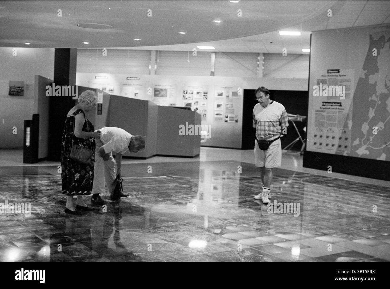 Schipholscope, Whizgle News, Dutch Desk, pays-Bas, 1950 - 2000 le 28-07-1994. L'image montre ces rubriques. La scène se déroule dans un intérieur spacieux, caractérisé par un design moderne et des surfaces élégantes. Le sol présente une finition en marbre poli réfléchissant qui brille sous l'éclairage ambiant. Différentes nuances de gris et des notes de blanc créent un motif subtil mais élégant sur le sol. Trois individus occupent l'espace. À gauche, une femme plus âgée aux cheveux blancs bouclés est habillée d'une longue robe à motifs qui contraste avec la tenue plus décontractée de son compagnon. Elle tient une petite poignée Banque D'Images