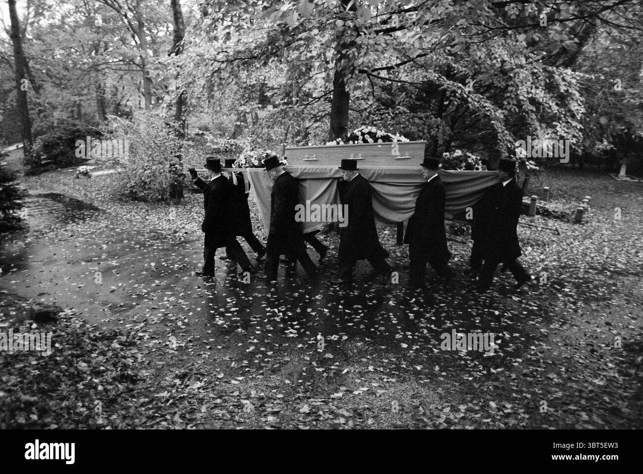 Conseiller funéraire Erftemeijer, Whizgle News, Dutch Desk, pays-Bas, 1950 - 2000 on 28-10-1994. Ces rubriques apparaissent dans l'image. Une scène sombre se déroule dans un cadre extérieur, caractérisé par une dense canopée d'arbres dont le riche feuillage fait allusion à la saison d'automne. Des feuilles, dans différentes nuances de brun, jaune et orange, tapissent le sol, créant un contraste frappant avec les vêtements sombres et discrets des personnages qui se déplacent à travers la scène. Six individus vêtus de longs manteaux noirs et de hauts chapeaux portent un grand cercueil orné, enveloppé par un tissu fluide qui adoucit sa présence imposante. Thei Banque D'Images