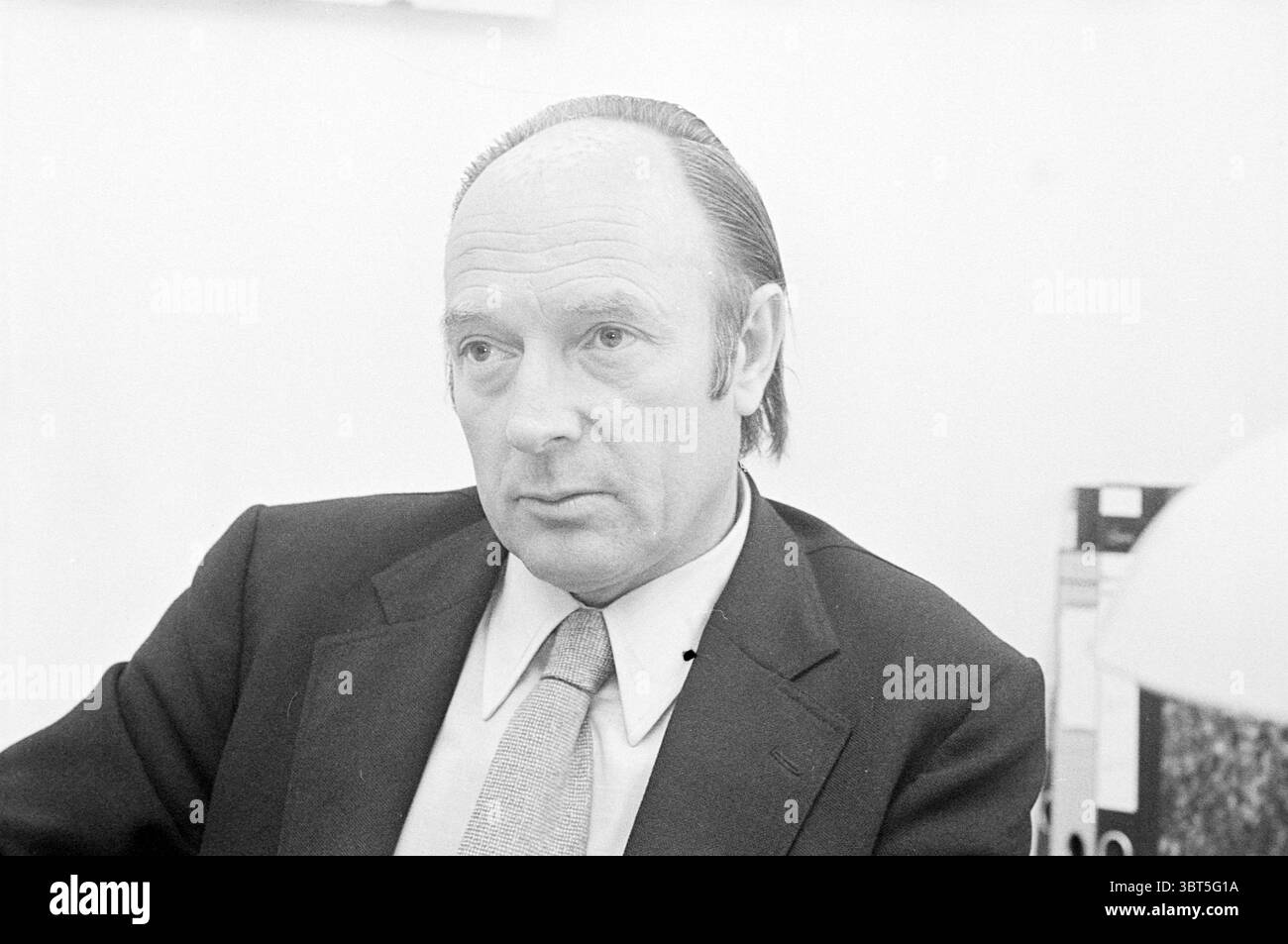 Portrait Mr. Möger Portraits, Whizgle News, Dutch Desk, pays-Bas, 1950 - 2000 le 21-09-1973. Ces rubriques sont illustrées dans l'image. Dans la scène, un homme est assis à un bureau, son expression réfléchie et sérieuse. Il a les cheveux reculés, soigneusement peigné le dos, et porte un costume sombre associé à une cravate de couleur claire, suggérant un cadre formel ou professionnel. L'arrière-plan est minimal, avec un mur neutre qui améliore l'accent sur lui. L’éclairage est doux mais direct, projetant de douces ombres qui mettent en valeur les contours de son visage et créent une atmosphère contemplative. Son regard est dirigé légèrement Banque D'Images