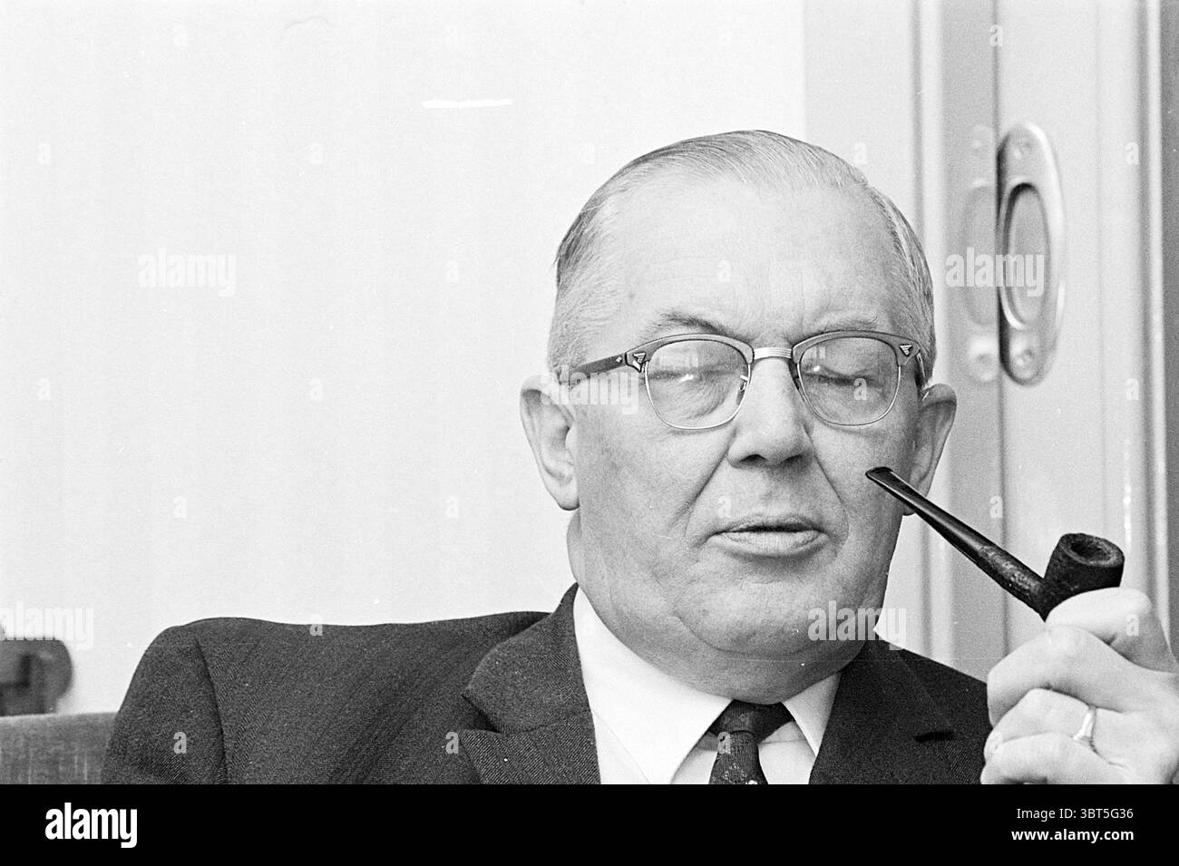 Portrait of a Man., Whizgle News, Dutch Desk, pays-Bas, 1950 - 2000. Ce sont les éléments de l'image. La scène met en scène un homme âgé assis confortablement, dégageant un sentiment de calme et de contemplation. Il a un teint clair et porte des cheveux gris soigneusement peignés, ainsi qu'une paire de lunettes à bords épais qui ajoutent à son apparence distinguée. Un costume sombre classique, conçu pour s'adapter, souligne son comportement formel. Il tient une pipe à ses lèvres, semblant savourer un moment de réflexion tranquille, avec un léger sourire évoquant ses pensées. L'arrière-plan se compose d'un mur doux et neutre, enh Banque D'Images