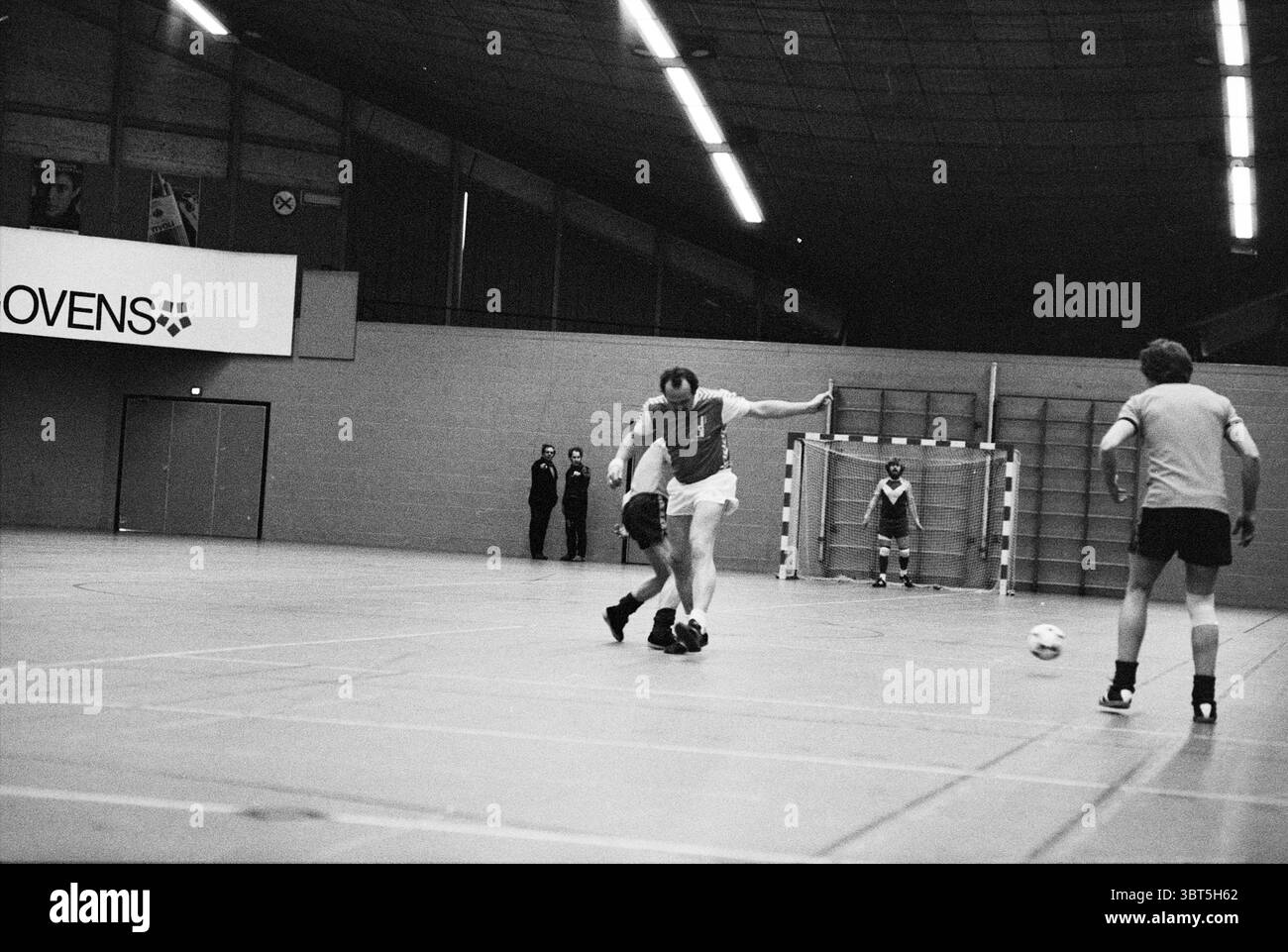 Carillon Boys - Wilms Floet Indoor football, Whizgle News, Dutch Desk, pays-Bas, 1950 - 2000 le 13-04-1981. Ces rubriques apparaissent dans l'image. La scène capture un match de football intérieur dynamique dans un gymnase vaste. Des plafonniers brillants baignent la zone dans une forte fluorescence, accentuant les sols lisses et polis qui reflètent l'énergie du jeu. À l'avant-garde, deux joueurs sont engagés dans un moment tendu ; l'un, portant un maillot rouge, semble être dans une position défensive alors qu'il tente d'intercepter le ballon, qui est une sphère blanche et dure roulant sur le sol. L'autre jeu Banque D'Images