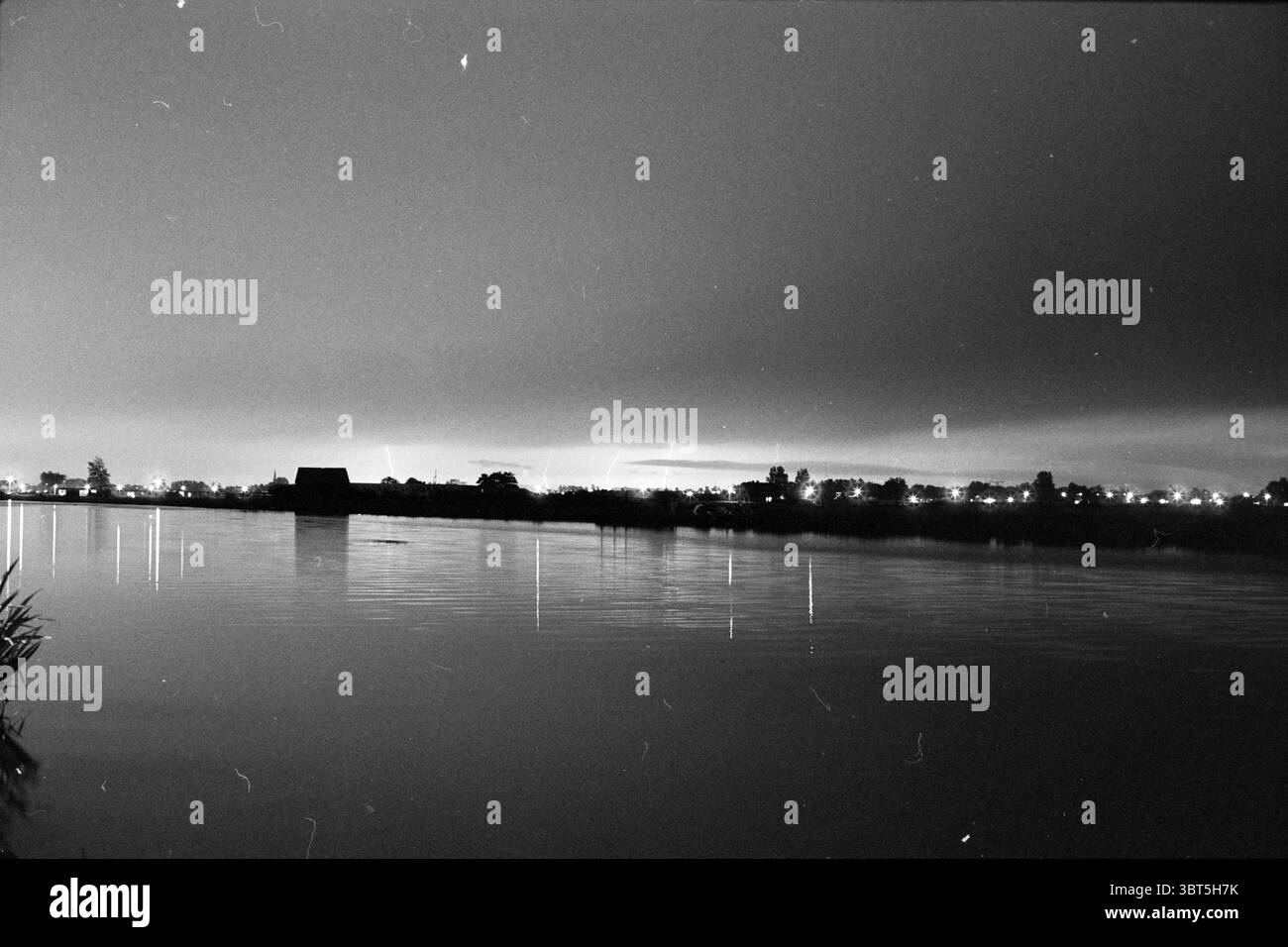 Lightning over H'gom Hillegom, Whizgle News, Dutch Desk, pays-Bas, 1950 - 2000 on 11-07-1991. Voici les rubriques de l'image. La scène présente un plan d'eau serein, reflétant un ciel sombre et silencieux entrecoupé d'une lumière douce et diffuse. L'horizon est légèrement éclairé, suggérant la présence de lumières de la ville au loin, créant un contraste doux avec l'ambiance autrement sombre. Silhouettés contre l'eau sont les contours de structures, peut-être des bâtiments ou une simple grange, qui ajoutent un élément de profondeur à la scène. Les formes sont sombres et indéfinies, contrastant nettement w Banque D'Images
