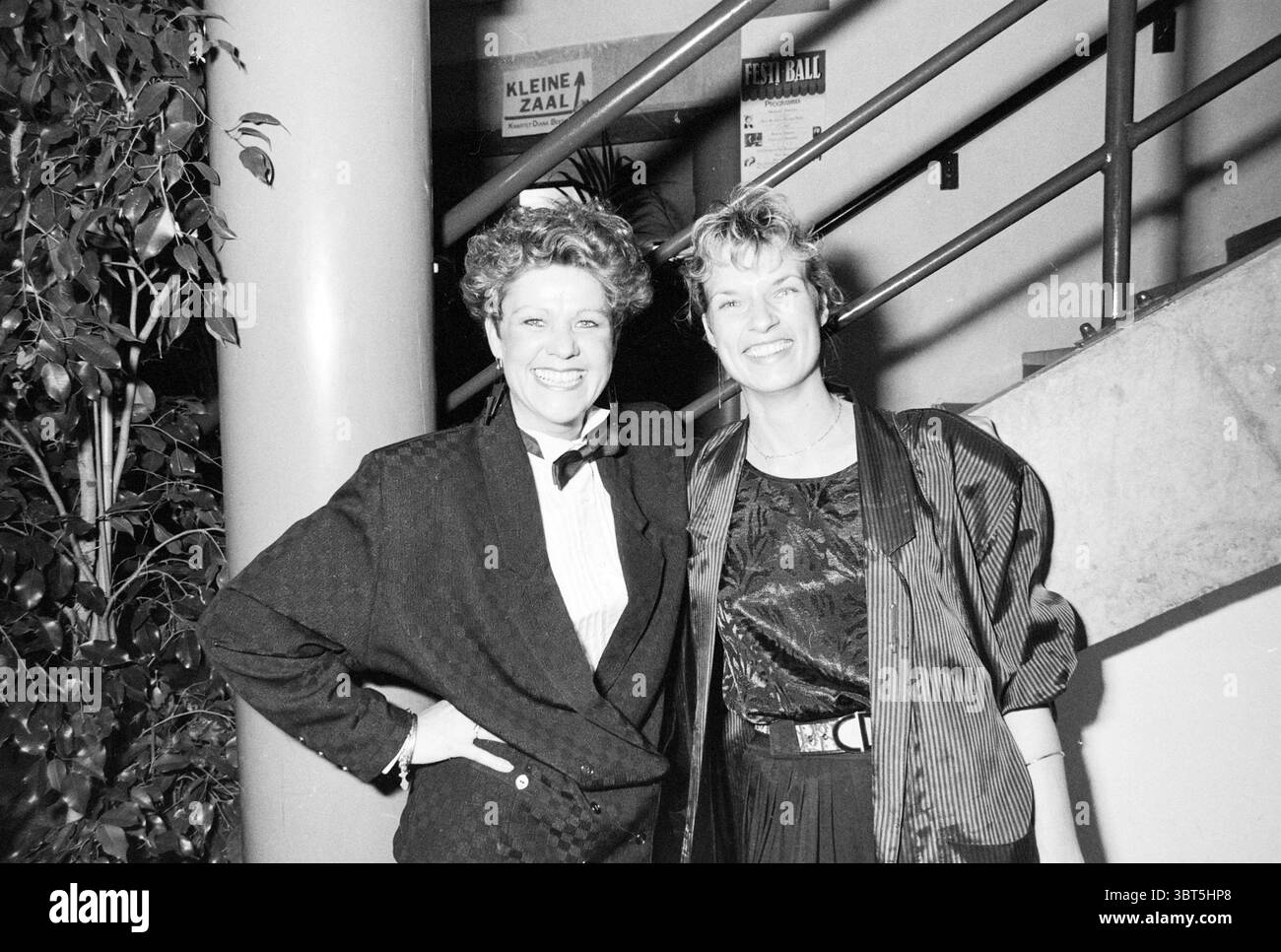 Festiball Concertgebouw Football Haarlem, Whizgle News, Dutch Desk, pays-Bas, 1950 - 2000 le 28-01-1989. Voici les rubriques de l'image. Dans la scène, deux femmes se tiennent étroitement ensemble, dégageant toutes deux un sentiment de confiance et de camaraderie. La toile de fond présente un escalier qui courbe autour, suggérant un cadre intérieur animé, éventuellement un événement ou un espace de rassemblement. Une femme sur la gauche a une coiffure distincte, caractérisée par des boucles volumineuses, qui ajoute une ambiance ludique mais sophistiquée. Elle est habillée dans une tenue sombre sur mesure avec un noeud papillon, dégageant un chic, presque ret Banque D'Images