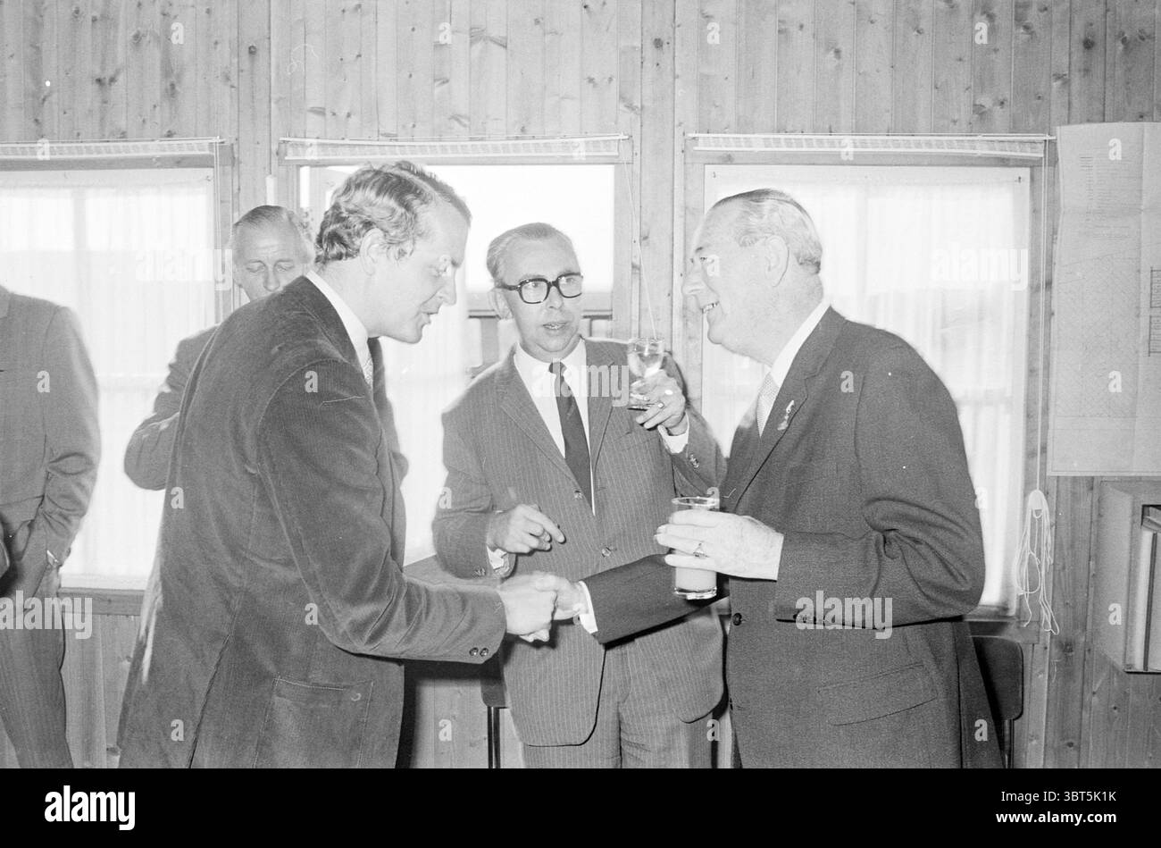 1ère pose de pierre bej. Home First Stone, Whizgle News, Dutch Desk, pays-Bas, 1950 - 2000 le 24-09-1971. L'image contient ces rubriques. La scène capture un rassemblement à l'intérieur, caractérisé par une atmosphère chaleureuse et accueillante. Les murs sont construits en bois clair, créant une toile de fond rustique et confortable. La lumière naturelle filtre à travers de grandes fenêtres, améliorant l'ambiance intime du cadre. Au premier plan, deux hommes bien habillés sont engagés dans la conversation. Un homme, portant un blazer foncé avec une texture velours, étend sa main dans un geste de salutation ou d'introduction. L'autre homme, habillé Banque D'Images