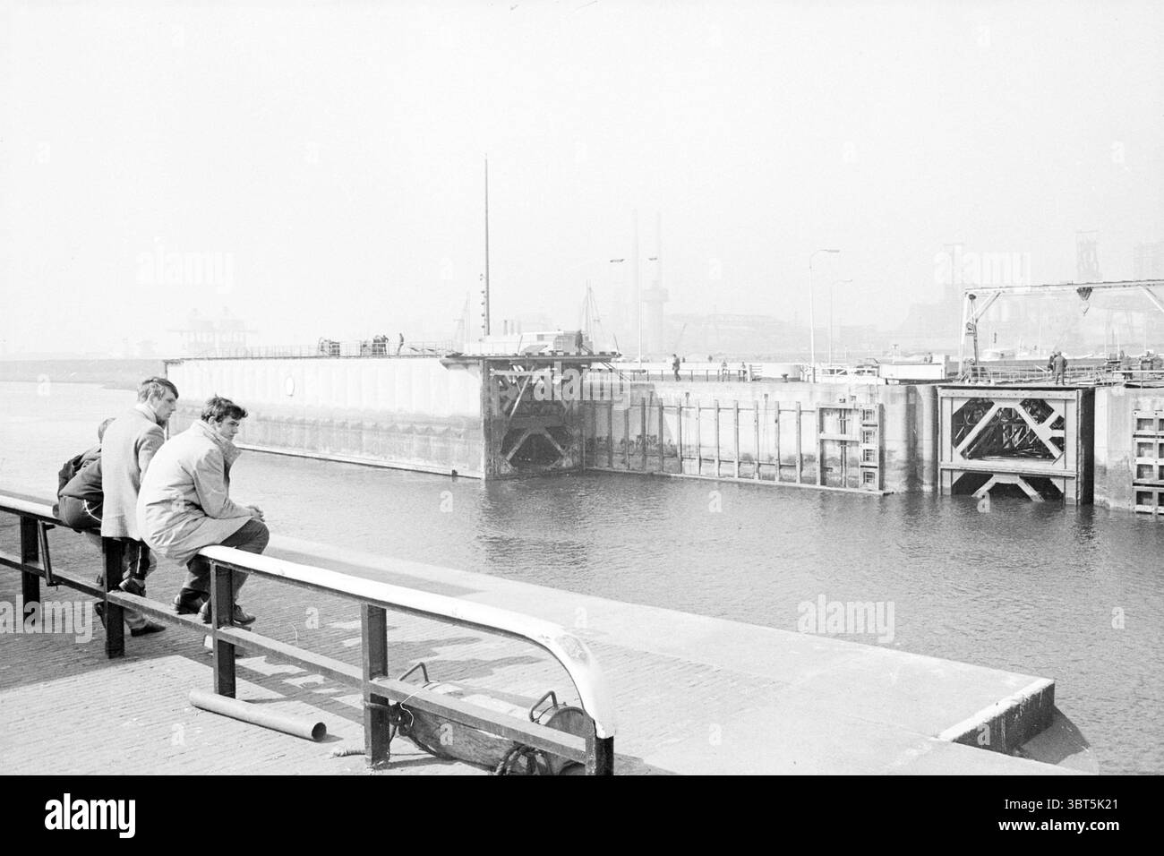 Les portes de verrouillage mobiles verrouillent les verrous de construction travaux hydrauliques, Whizgle News, Dutch Desk, pays-Bas, 1950 - 2000 sur 14-04-1971. Ces rubriques sont illustrées dans l'image. La scène capture un front de mer serein, où l'eau tranquille reflète des tons doux et discrets de gris et de bleu, suggérant une atmosphère calme. Au premier plan, une balustrade basse court parallèlement au bord de l'eau, sa peinture blanche légèrement altérée, indiquant des années d'exposition aux éléments. Deux personnages, assis sur la balustrade, semblent contemplatifs alors qu'ils regardent au-dessus de l'eau. Leurs vêtements sont décontractés, et les teintes discrètes de leur atti Banque D'Images