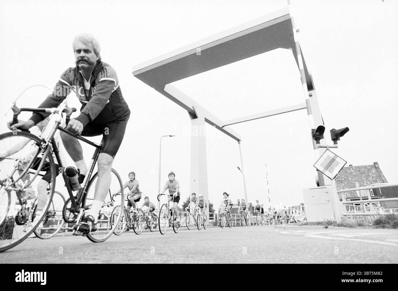 Ronde de moustiques au pont Spaarndam Muggeronde, Whizgle News, Dutch Desk, pays-Bas, 1950 - 2000 le 27-07-1986. L'image inclut ces rubriques. La scène présente un événement cycliste dynamique sur un ciel couvert et sombre qui projette une lumière silencieuse sur le paysage. Un groupe de cyclistes se déplace en ligne le long d'une route pavée, leurs vélos variant dans la conception mais principalement élégant et léger, adapté à la course. Au premier plan, un cycliste se démarque avec une moustache distinctive, portant un maillot de cyclisme sombre et un short. Son expression dévouée et sa posture concentrée suggèrent qu'il est Banque D'Images