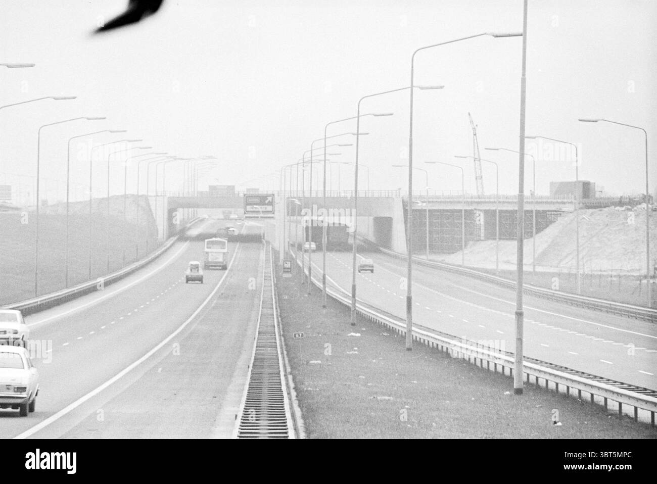 Schiphol Railway line Rijksweg 4 chemins de fer Schiphol Schiphol Rijksweg 4, Whizgle News, Dutch Desk, pays-Bas, 1950 - 2000 on 23-12-1975. Ce sont les éléments de l'image. La scène représente une autoroute large et ouverte s'étendant au loin, divisée en deux voies avec un séparateur visible séparant les véhicules circulant dans des directions opposées. La route semble bien entretenue mais est relativement peu fréquentée, ce qui donne un sentiment d'ouverture. Des véhicules sont présents, y compris un couple de voitures et un camion plus grand, qui sont positionnés le long des voies, soulignant l'autoroute un peu désolée à Banque D'Images
