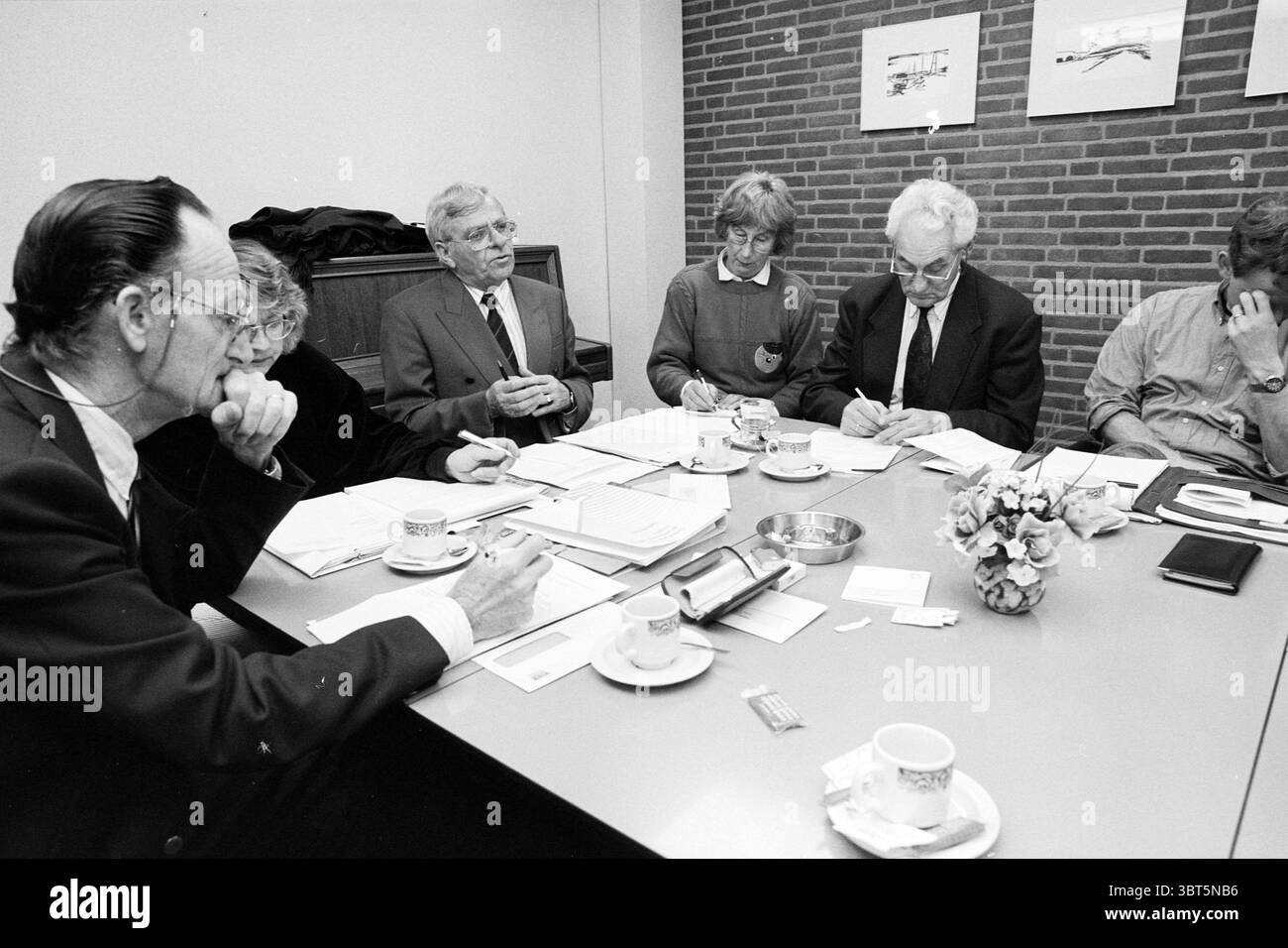 Réunion du conseil du village de Zwanenburg Zwanenburg, Whizgle News, Dutch Desk, pays-Bas, 1950 - 2000 le 12-04-1995. Voici les rubriques de l'image. Un groupe de six personnes est assis autour d'une table rectangulaire, engagé dans une discussion. Le cadre offre une atmosphère chaleureuse et accueillante, mise en valeur par un éclairage ambiant doux qui illumine la pièce. Les murs sont ornés de briques de couleur claire, donnant à l'environnement une sensation confortable et professionnelle. La table est encombrée de divers documents, cahiers et stylos, indiquant une réunion de travail. De petites tasses et soucoupes élégantes sont placées dedans Banque D'Images