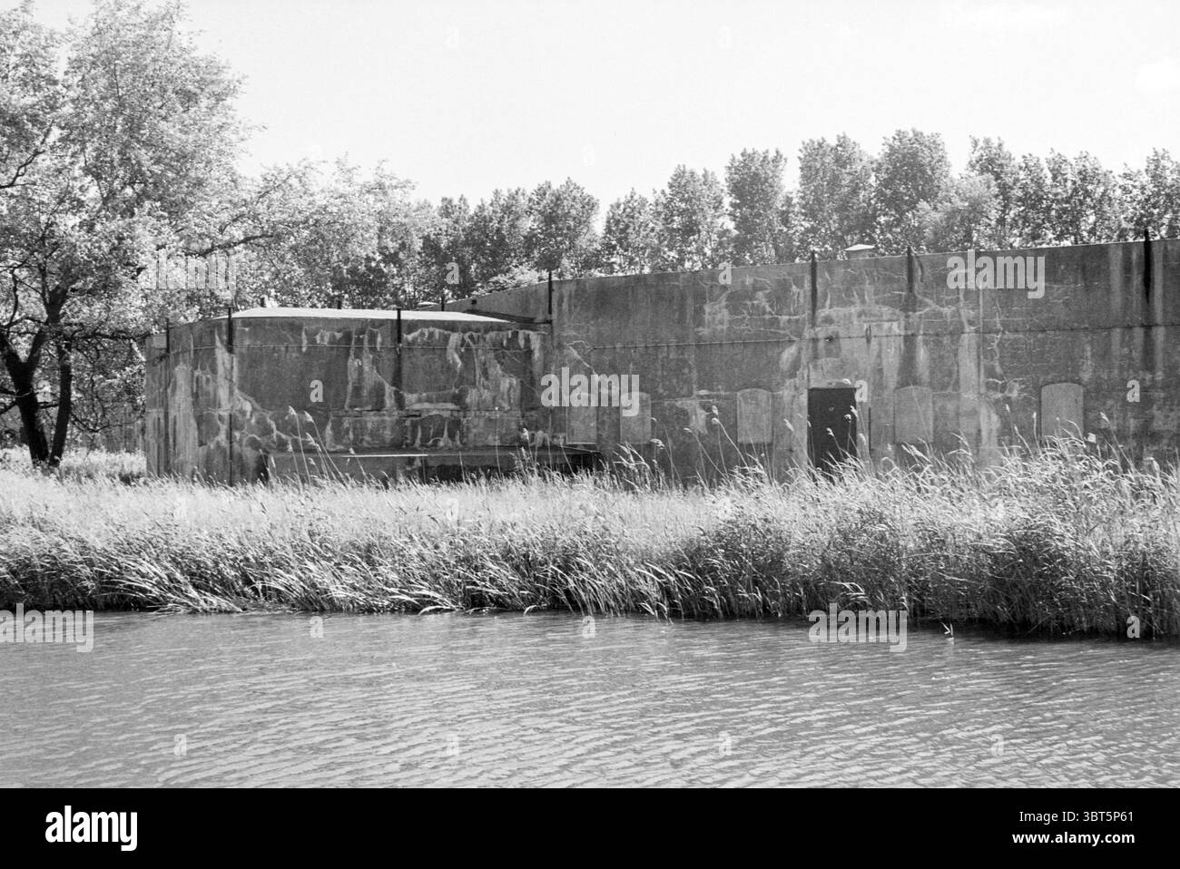 Fort Penningsveer Haarlemmerliede Penningsveer, Whizgle News, Dutch Desk, pays-Bas, 1950 - 2000 le 22-05-1990. L'image montre ces rubriques. La scène représente une structure austère et altérée située près d'un plan d'eau. Le bâtiment, caractérisé par sa surface rugueuse et grise en béton, révèle des signes d'âge avec des taches de peinture et de texture décolorées qui racontent des histoires de son passé. La structure est quelque peu rectangulaire et à sommet plat, s'élevant en évidence au milieu d'une toile de fond naturelle. Entourant le bâtiment, une mer de hautes herbes oscille doucement dans une brise légère, leurs nuances variées de vert et de Go Banque D'Images