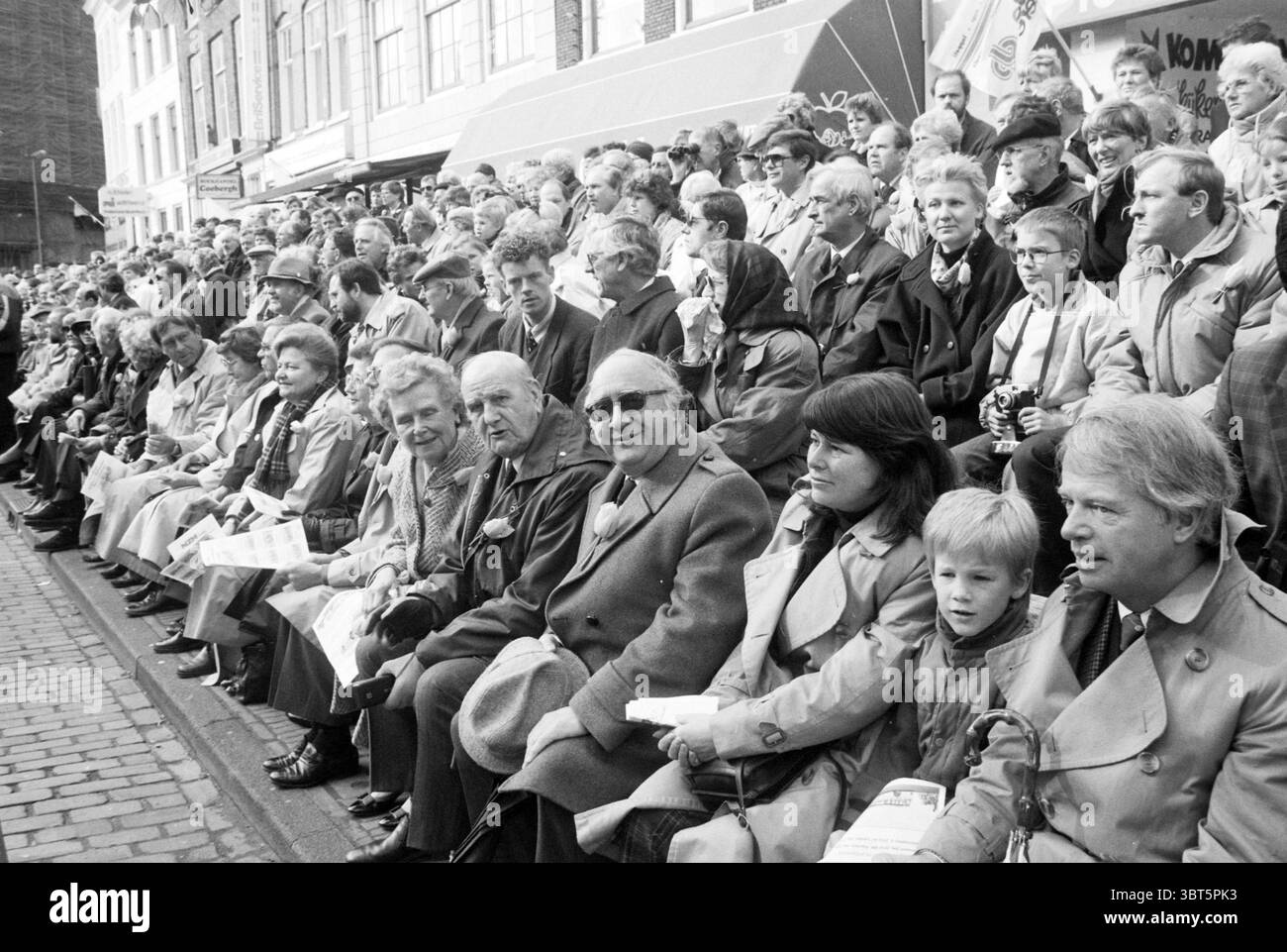 Audience à Bloemencorso Haarlem - Hillegom, Whizgle News, Dutch Desk, pays-Bas, 1950 - 2000 le 22-04-1989. L'image inclut ces rubriques. La scène représente un grand rassemblement de personnes assis étroitement ensemble, créant un sentiment de communauté et d'anticipation. La foule est principalement habillée de manteaux de couleur plus claire, suggérant une saison douce ou transitoire, peut-être à la fin du printemps ou au début de l'automne. Les individus varient en âge, des hommes et des femmes âgés aux enfants, indiquant un événement multigénérationnel. L'ambiance est vive et engagée, car beaucoup de gens semblent souriants ou mécontents Banque D'Images