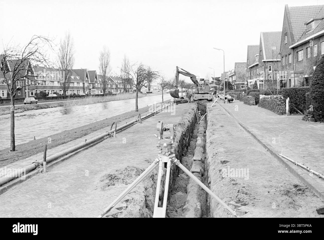 Usines d'assainissement Asterkade usines d'assainissement Heemstede Asterkade pays-Bas, Whizgle News, Dutch Desk, pays-Bas, 1950 - 2000 on 11-02-1977. L'image inclut ces rubriques. La scène présente une longue tranchée rectangulaire creusée le long d'une rue, bordée d'un mélange de bâtiments résidentiels et d'un canal. La tranchée est bien en évidence, avec son sol terreux exposé contrastant avec le trottoir pavé. Le long de la tranchée, de la machinerie lourde, y compris une excavatrice, est positionnée, ce qui suggère des travaux de construction ou de réparation en cours. De chaque côté de la tranchée, les maisons présentent une variété de A. Banque D'Images