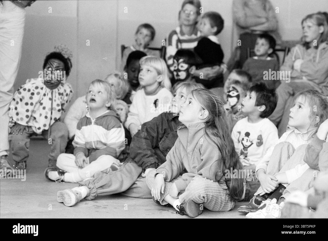 Fête des enfants dans Zero Parties, Whizgle News, Dutch Desk, pays-Bas, 1950 - 2000 le 05-05-1988. Ces rubriques sont illustrées dans l'image. Dans un cadre intérieur animé, un groupe diversifié d'enfants est assis sur le sol, encadré en demi-cercle. Leurs expressions sont un mélange d'admiration et de curiosité, tous les yeux dirigés vers une figure ou une source de divertissement juste hors de vue. Les enfants sont habillés de vêtements décontractés et colorés. Certains portent des vestes brillantes, tandis que d'autres sont en chandails confortables ou des chemises ludiques représentant des personnages de dessins animés. Un élément notable est un enfant avec de longs cheveux bruns, étroitement wa Banque D'Images
