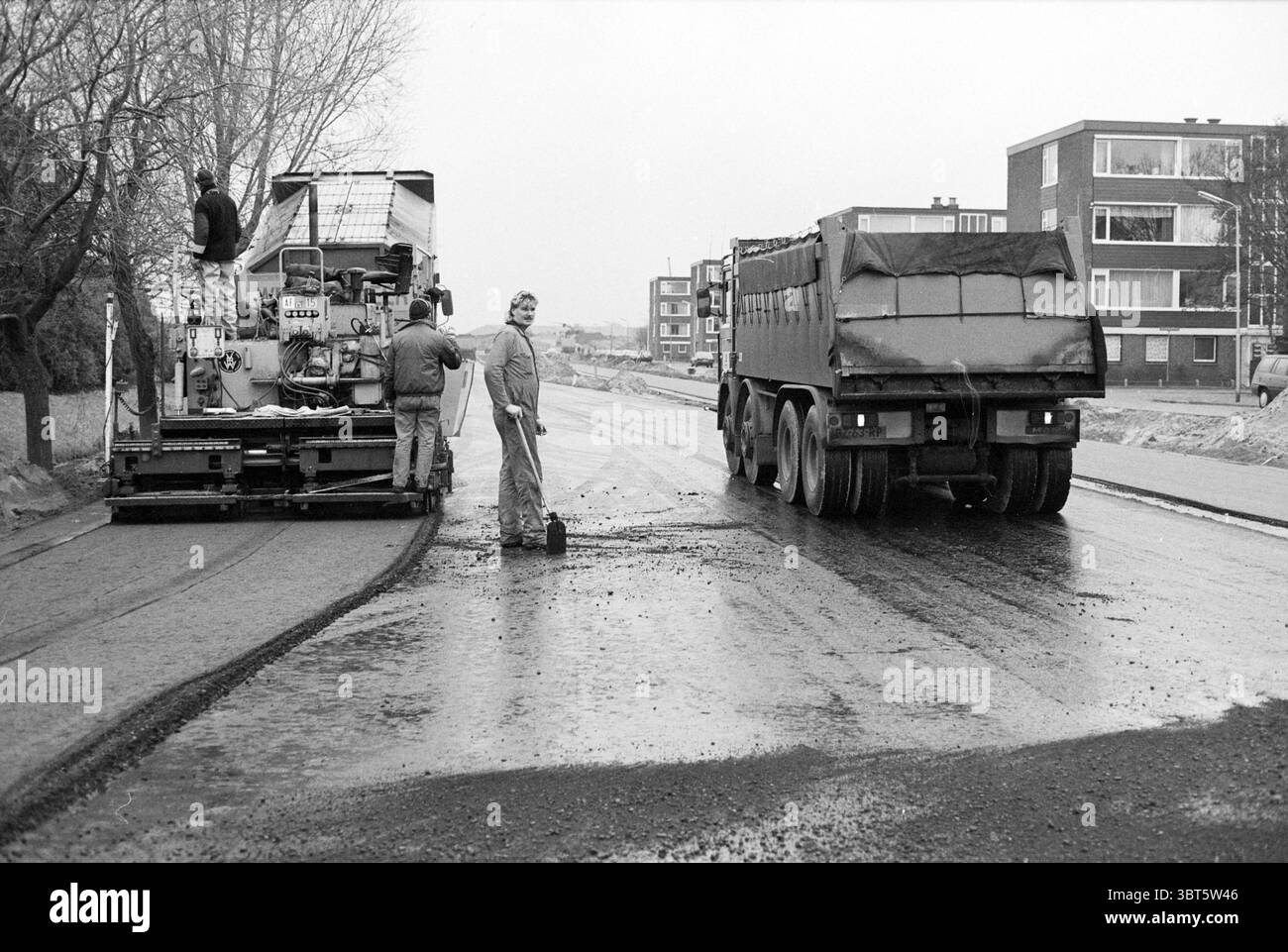 Road construction., Whizgle News, Dutch Desk, pays-Bas, 1950 - 2000. L'image contient ces rubriques. La scène représente une journée grise et nuageuse dans un environnement urbain, caractérisé par un cadre industriel. Au premier plan, deux personnes travaillent à proximité d'une grosse machine, possiblement liée à la construction ou à l'entretien de routes. Un ouvrier, vêtu de vêtements sombres et amples, se tient debout, son corps légèrement tourné vers le spectateur, tandis que l'autre se penche sur la machine, l'ajustant apparemment ou vérifiant sa fonctionnalité. La machinerie elle-même est encombrante et Banque D'Images