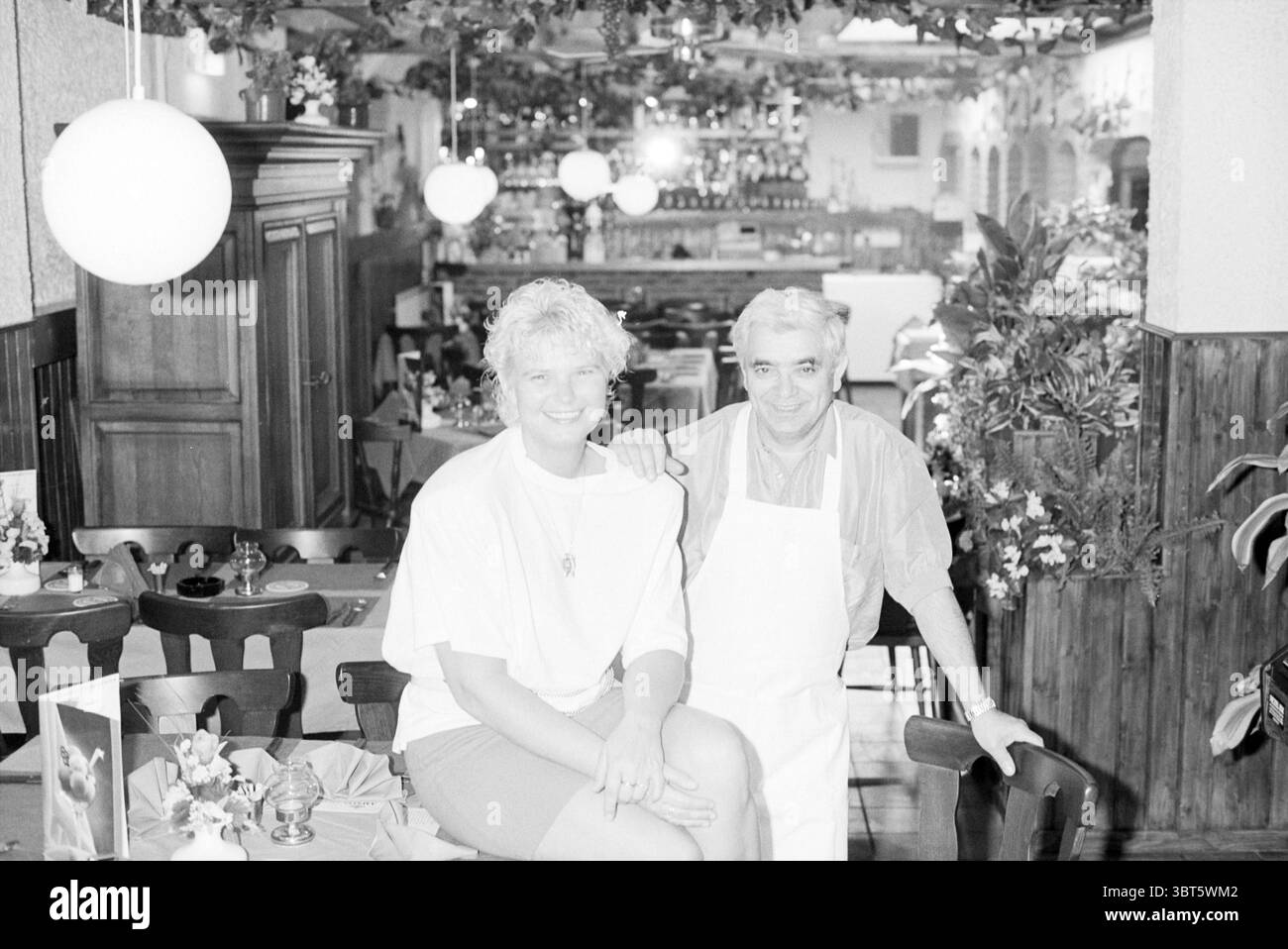 Restaurant italien IJmuiden IJmuiden pays-Bas, Whizgle News, Dutch Desk, pays-Bas, 1950 - 2000 le 19-05-1992. Ces rubriques apparaissent dans l'image. La scène capture un intérieur de restaurant confortable, caractérisé par une ambiance accueillante. Une femme est assise sur le bord d'une table en bois, vêtue d'une chemise blanche ample et d'un short, ses cheveux courts et bouclés contrastant avec sa tenue lumineuse. Elle a une expression joyeuse, rayonnant de chaleur et de convivialité. À côté d'elle se tient un homme portant un tablier de couleur claire au-dessus d'une chemise à col, présentant une position accueillante. Ses cheveux légèrement grisonnés et Banque D'Images