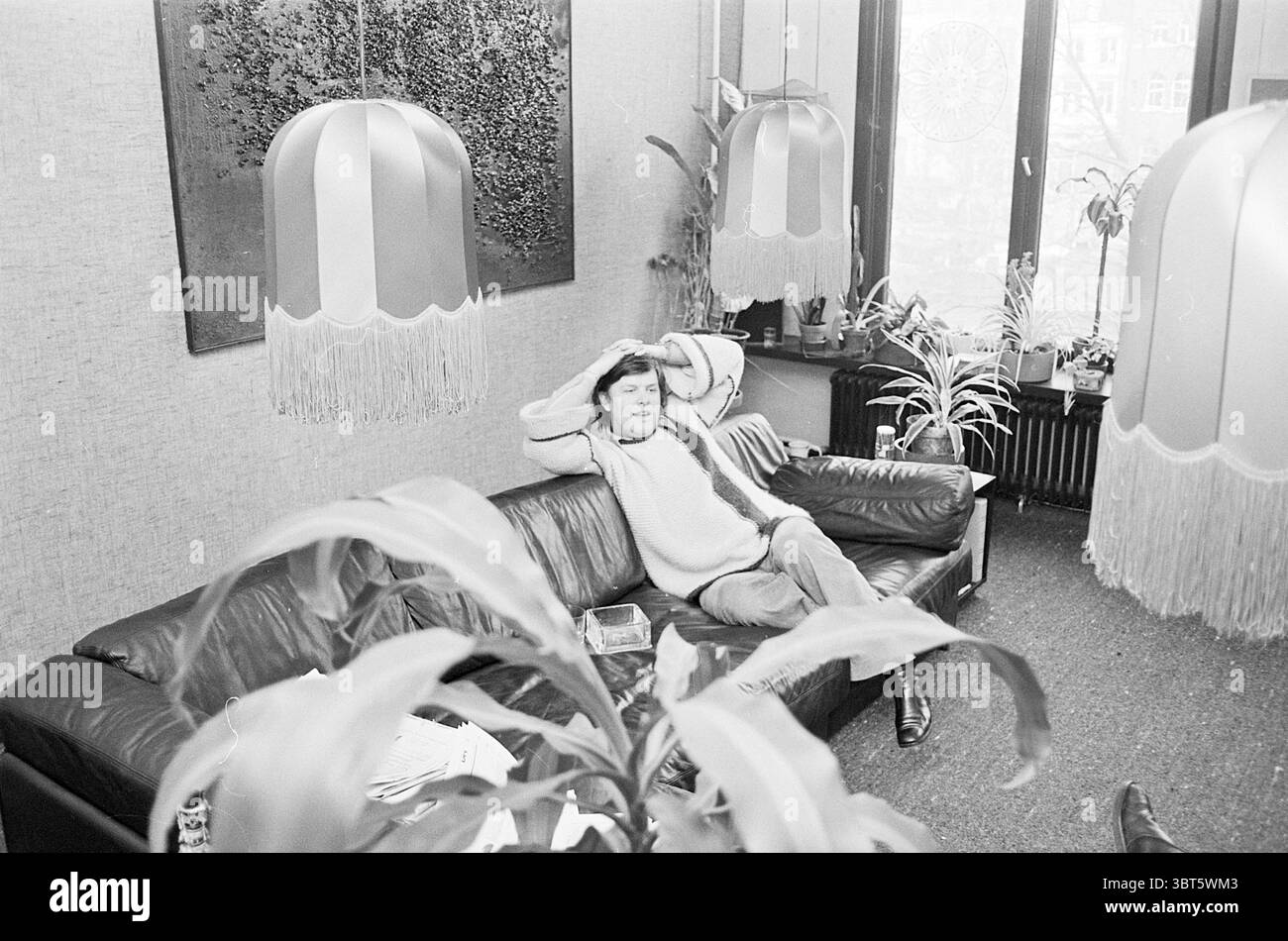 Man on Couch, Whizgle News, Dutch Desk, pays-Bas, 1950 - 2000 en 1976. Ce sont les éléments de l'image. Dans un espace intérieur confortable, une personne est assise confortablement sur un canapé en cuir foncé, se penchant en arrière avec les mains derrière la tête, exsudant une posture détendue. L'individu est habillé de façon décontractée, suggérant une atmosphère décontractée. Autour d'eux se trouvent quelques plantes, ajoutant une touche de verdure et de vie à la scène. Notons les lampes suspendues élégantes, avec des nuances de tissu dans des tons doux de couleurs pastel, ornés de franges délicates qui balancent doucement. Les murs sont revêtus d'un materia beige doux Banque D'Images