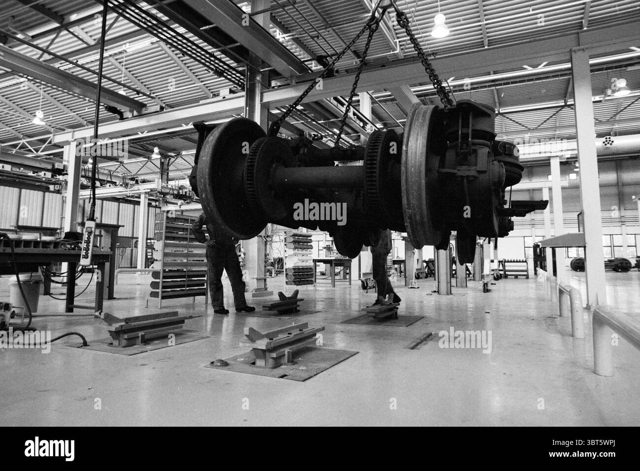 NS main Workshop bogie Department Railways, Whizgle News, Dutch Desk, pays-Bas, 1950 - 2000 le 05-04-1990. L'image contient ces rubriques. La scène capture un espace de travail industriel caractérisé par un grand composant de machinerie lourd déplacé en position. L'environnement est vaste avec de hauts plafonds, accentués par des lumières fluorescentes aériennes qui projettent un éclairage lumineux et uniforme sur toute la zone. L'objet dominant est un ensemble volumineux, probablement une partie d'un moteur ou d'un mécanisme similaire, suspendu à une chaîne robuste. La machinerie présente des détails complexes, y compris des engrenages et métalliques Banque D'Images