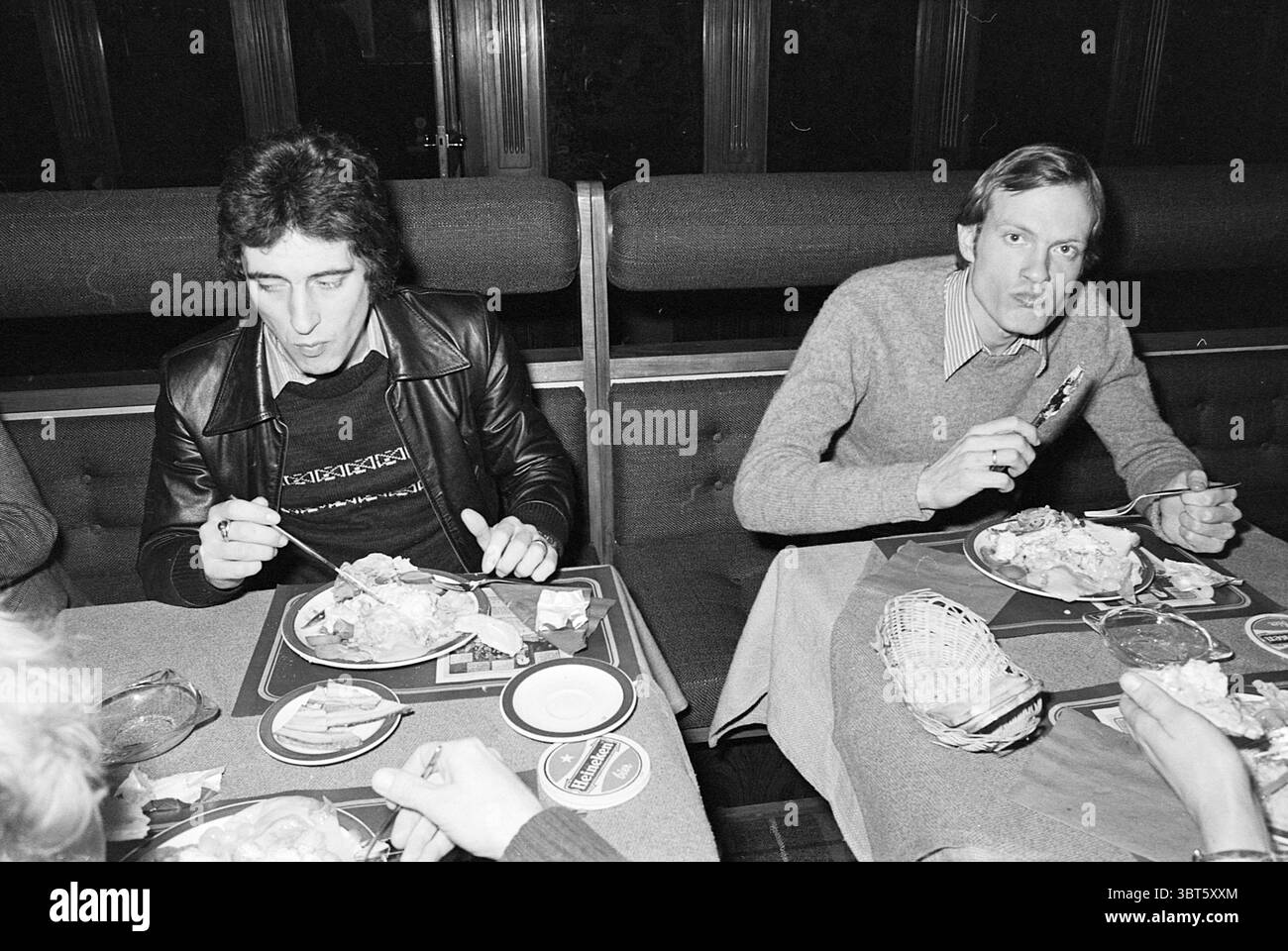 Quelques hommes ayant un repas commun., Whizgle News, Dutch Desk, pays-Bas, 1950 - 2000. Ce sont les éléments de l'image. Dans un cadre de salle à manger faiblement éclairé, deux personnes s'assoient à une table recouverte d'un tissu à motifs subtils. L'atmosphère suggère une ambiance décontractée mais intime, renforcée par la lueur chaude des plafonniers qui créent un contraste doux avec l'environnement sombre. Sur la table, les assiettes sont remplies de repas copieux qui comprennent des éléments reconnaissables tels que du poulet rôti et des accompagnements, donnant une sensation de réconfort. Les condiments et les ustensiles sont soigneusement disposés, accentuation Banque D'Images