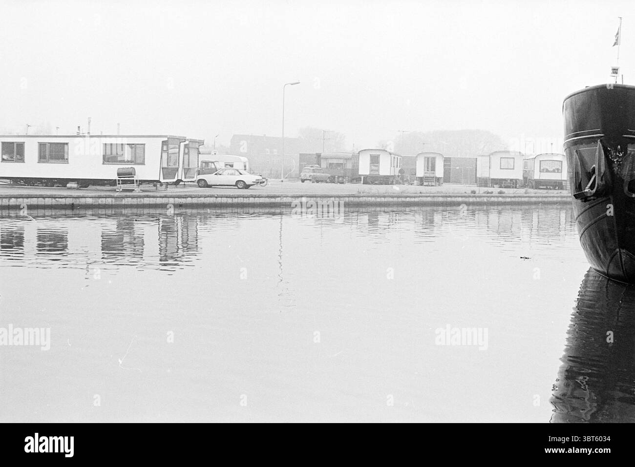 Vue d'ensemble des caravanes caravanes Heemstede caravanes caravanes caravanes résidants, Whizgle News, Dutch Desk, pays-Bas, 1950 - 2000 sur 27-02-1976. Voici les rubriques de l'image. La scène présente un cadre paisible au bord de l'eau enveloppé d'une lumière douce et silencieuse qui suggère une journée nuageuse. La surface de l’eau est lisse et miroir, reflétant les formes et les couleurs de l’environnement de manière sereine. Sur le côté droit, un grand bateau à coque sombre est positionné près du bord de l'eau, son arc courbant élégamment vers le spectateur. Sa surface brillante contraste avec les teintes douces du Banque D'Images