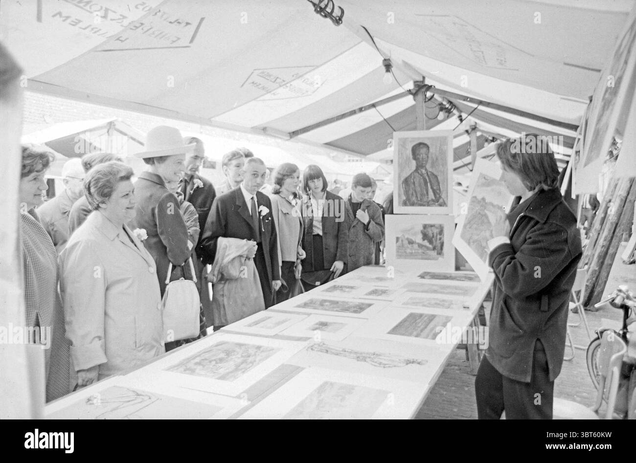 Art Market Market, Whizgle News, Dutch Desk, pays-Bas, 1950 - 2000 on 11-06-1965. Ces rubriques apparaissent dans l'image. La scène se déroule dans un cadre de marché animé, où une scène vibrante est capturée sous une grande verrière, partiellement ombragée du soleil. L'atmosphère est remplie d'un sentiment de curiosité et d'engagement parmi un groupe diversifié de personnes, toutes attirées vers une longue table ornée d'un éventail d'œuvres d'art. Au premier plan, un artiste se tient confiant, brandissant une œuvre d'art pour que tous puissent la voir, faisant des gestes animés comme s'il l'expliquait. Vêtue d'une veste sombre, l'artiste dégage un Banque D'Images