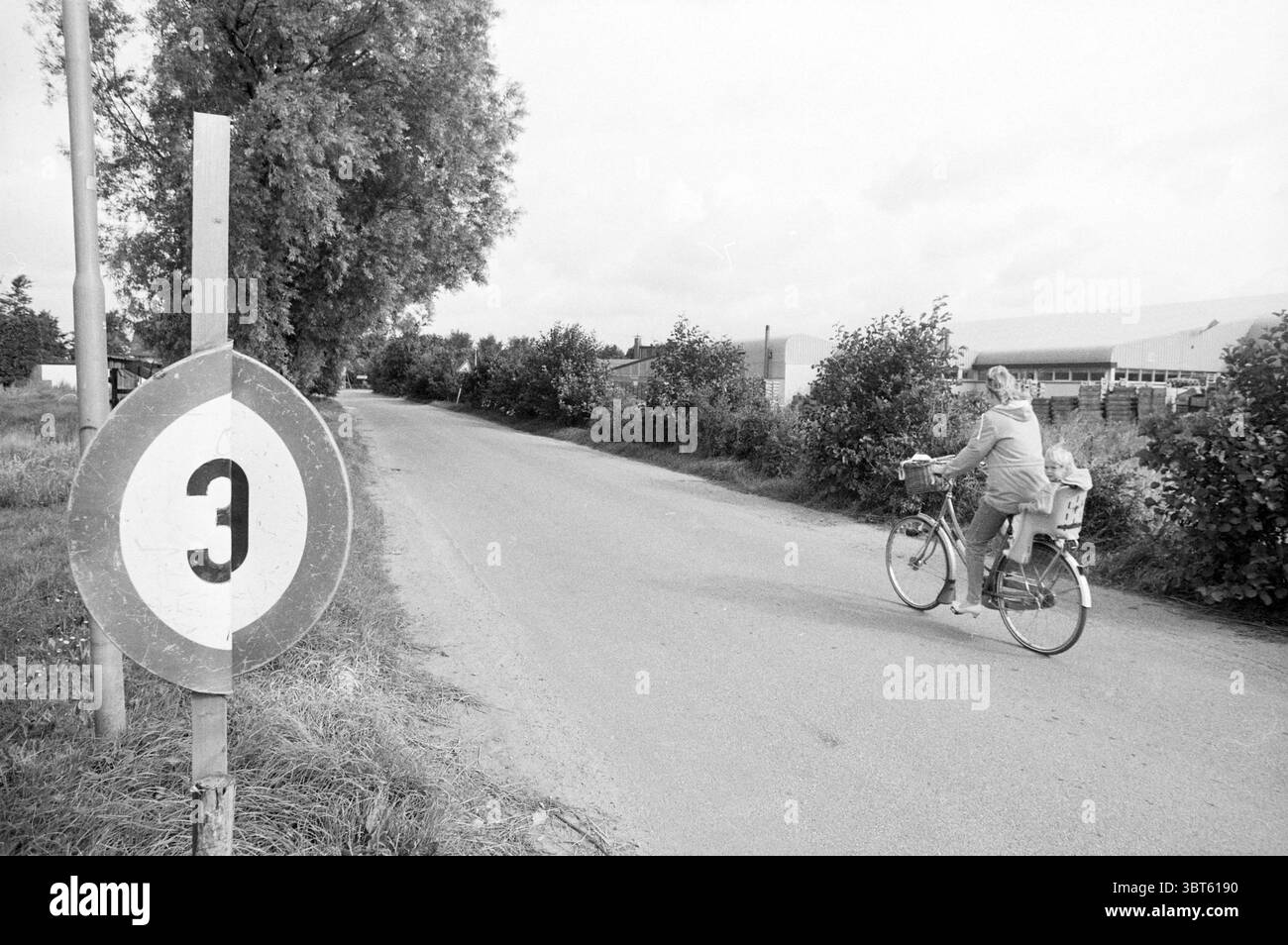 Panneau 3 km Hofgeesterweg Velserbroek Borden Velserbroek Hofgeesterweg, Whizgle News, Dutch Desk, pays-Bas, 1950 - 2000 on 01-08-1988. L'image contient ces rubriques. Dans cette scène, une route rurale tranquille s'étend au loin, flanquée de verdure luxuriante des deux côtés. Le cadre offre une atmosphère calme et sereine, favorisant un sentiment de tranquillité. À gauche, un grand panneau circulaire se tient bien en évidence, avec un chiffre noir gras '3' sur fond blanc, indiquant une distance ou une direction. Les bords rugueux du panneau suggèrent qu'il a été altéré par le temps et les éléments tout en maintenant Banque D'Images