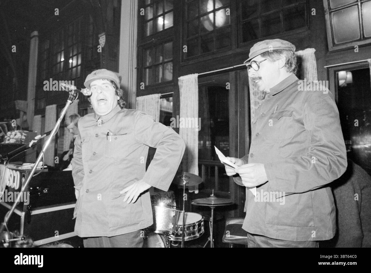 Udo achète restaurant de fête de mariage Haarlem station Haarlem pays-Bas, Whizgle News, Dutch Desk, pays-Bas, 1950 - 2000 le 24-11-1979. Ces rubriques apparaissent dans l'image. Dans une pièce faiblement éclairée, deux hommes sont le point focal, debout près d'un microphone et d'un petit ensemble d'instruments de musique, y compris une batterie sertie en arrière-plan. Le cadre semble être un espace de rassemblement ou de performance, caractérisé par de grandes fenêtres qui permettent à une lueur silencieuse de filtrer, suggérant que c’est le soir. Les hommes sont vêtus d'uniformes sombres, chacun portant une casquette de style militaire. Un homme apparaît animé, avec un w Banque D'Images